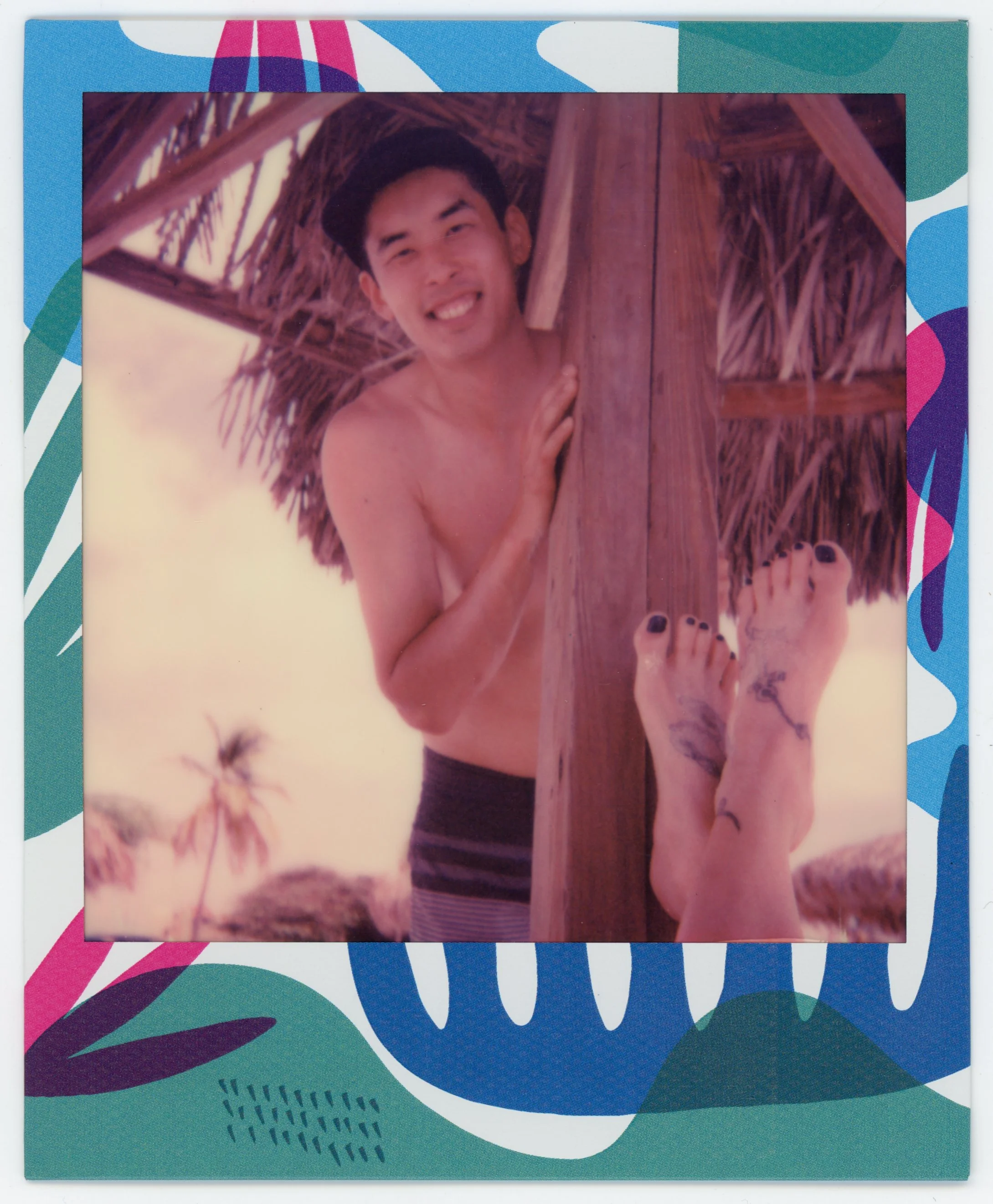 A young man with dark hair smiling and peeking out from behind a wooden post, with a thatched roof in the background. The photo is framed with colorful, abstract patterned border and shows tattoos on feet at the bottom of the image.