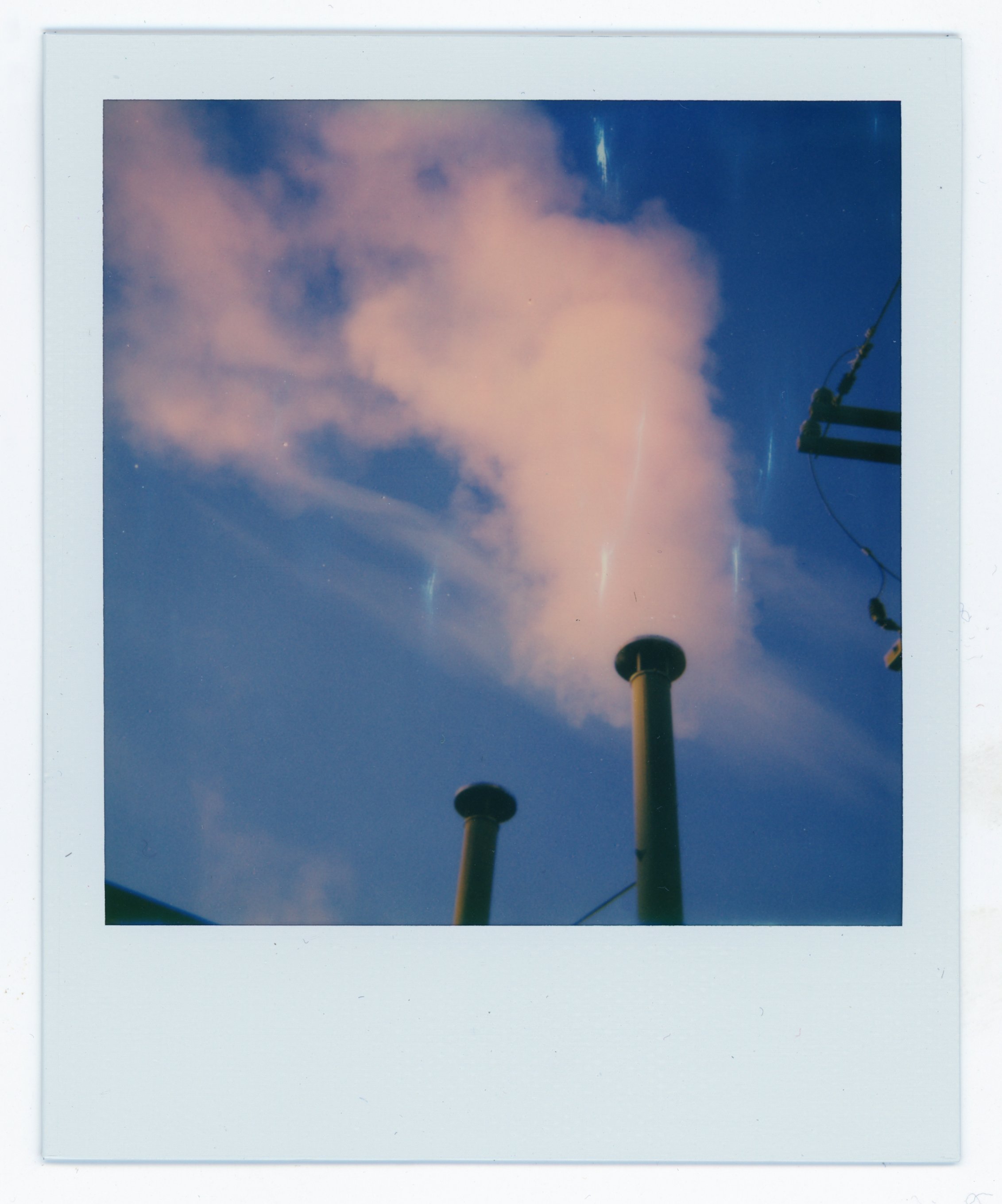 A Polaroid photo of clouds and sunset sky with two industrial chimneys and part of a utility pole with wires.