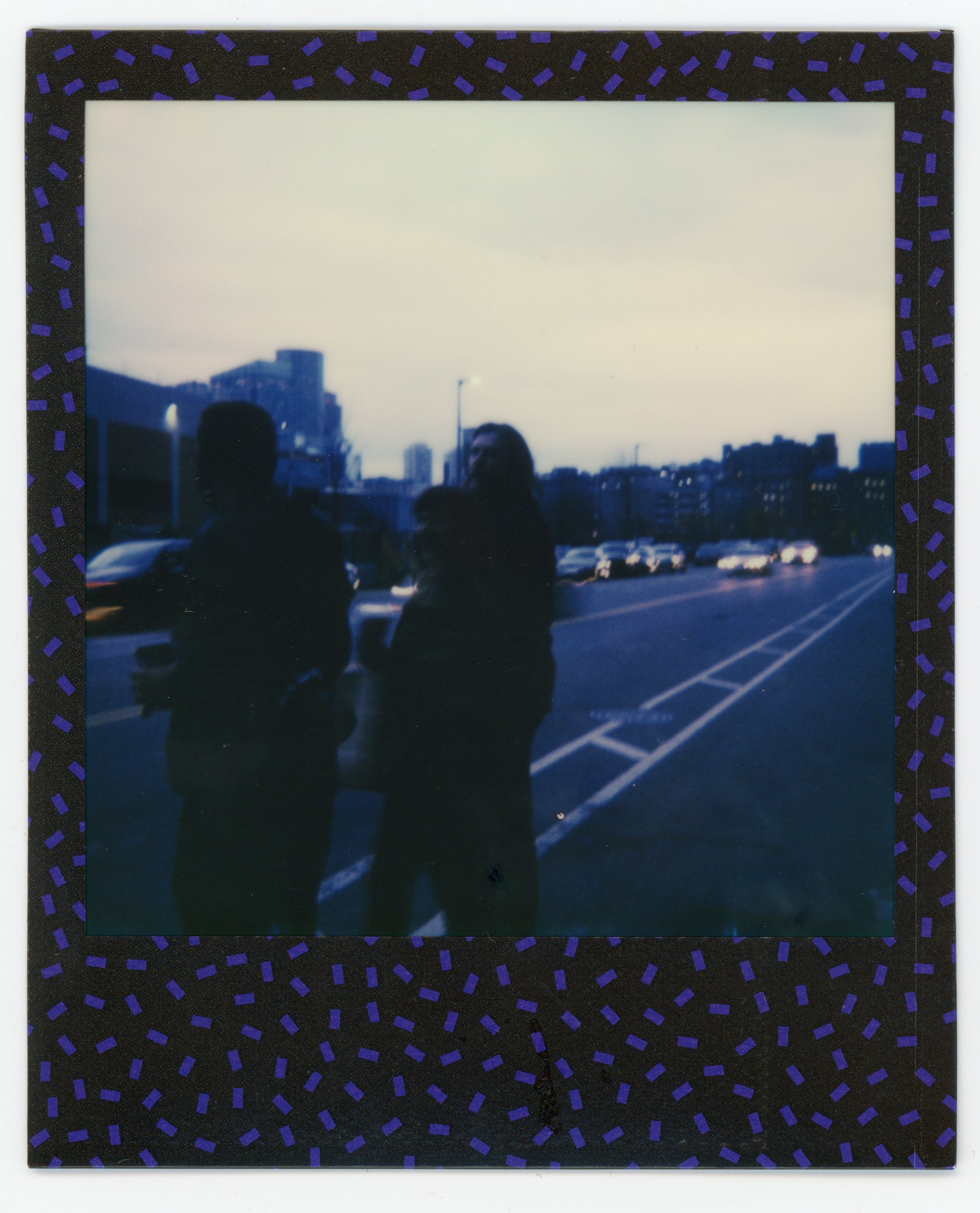 Two people standing on a city street during dusk, with cars driving by in the background and buildings visible in the distance.