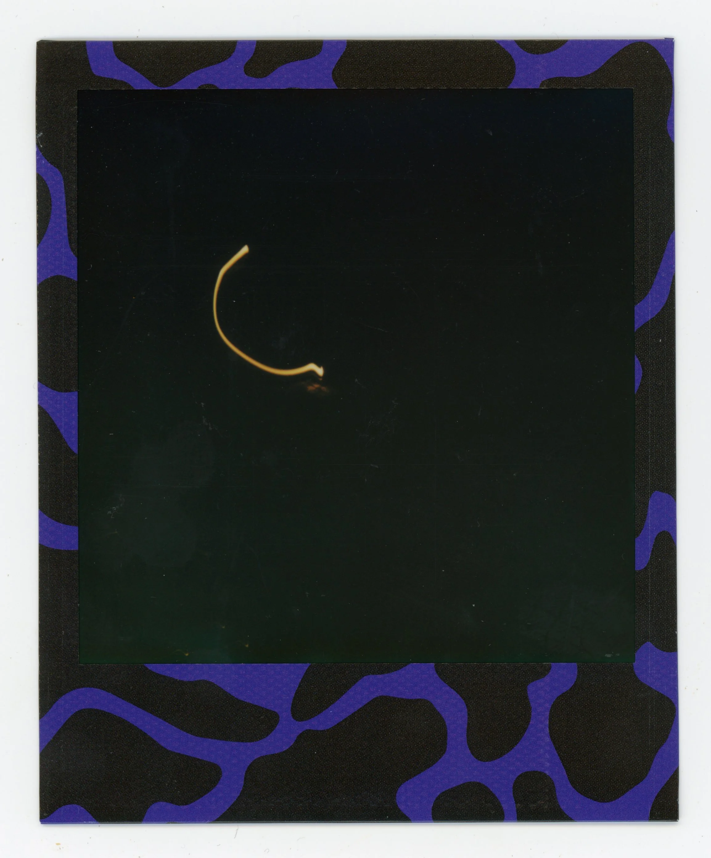 Dark Polaroid photo with a neon yellow object, possibly a light trail or filament, against a black background, in a purple and black patterned frame.