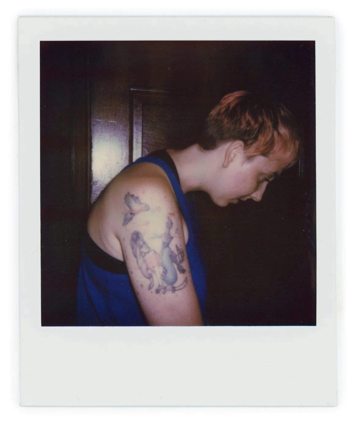 A person with short reddish hair, wearing a blue sleeveless shirt, showing a colorful tattoo of a dragon and other elements on their upper arm, with a dark wooden background.