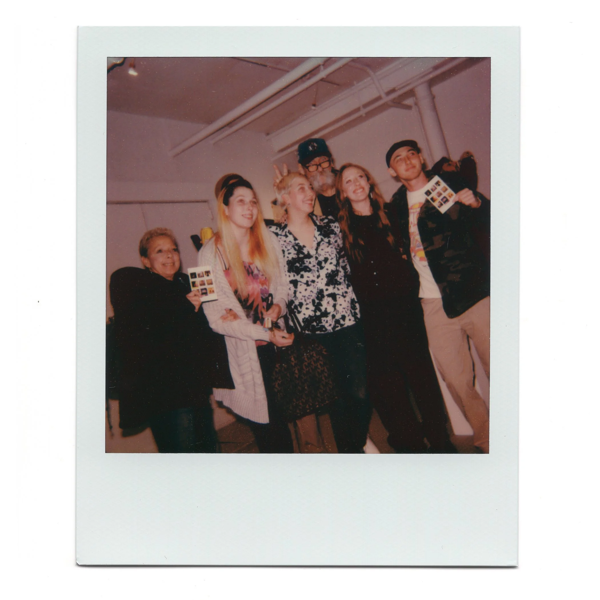 A group of five people, young and older, smiling and posing for a photo at an indoor event, with some holding small photo strips or cards, against a plain wall with ceiling pipes visible.