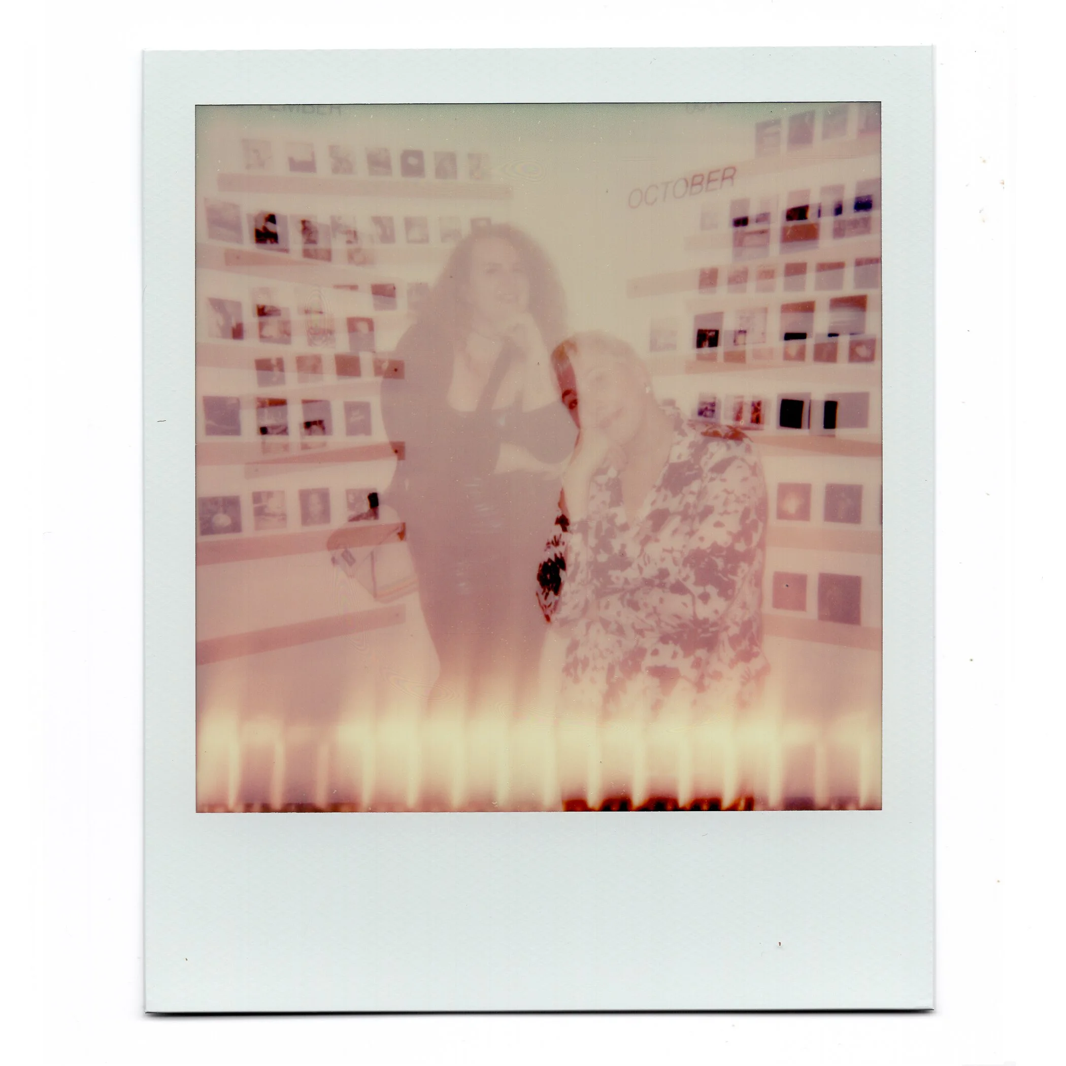 A faded polaroid photo of two women in front of a wall with photo displays, one standing and the other seated, with the word 'October' written on the wall.