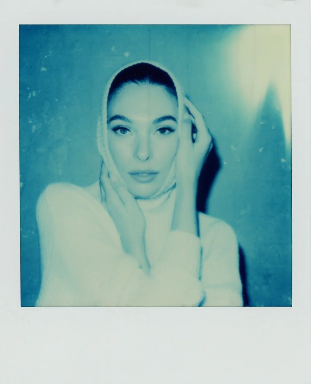 Polaroid photo of a woman with short hair, wearing a light-colored top, holding her face with her hands, and looking at the camera with a neutral expression.