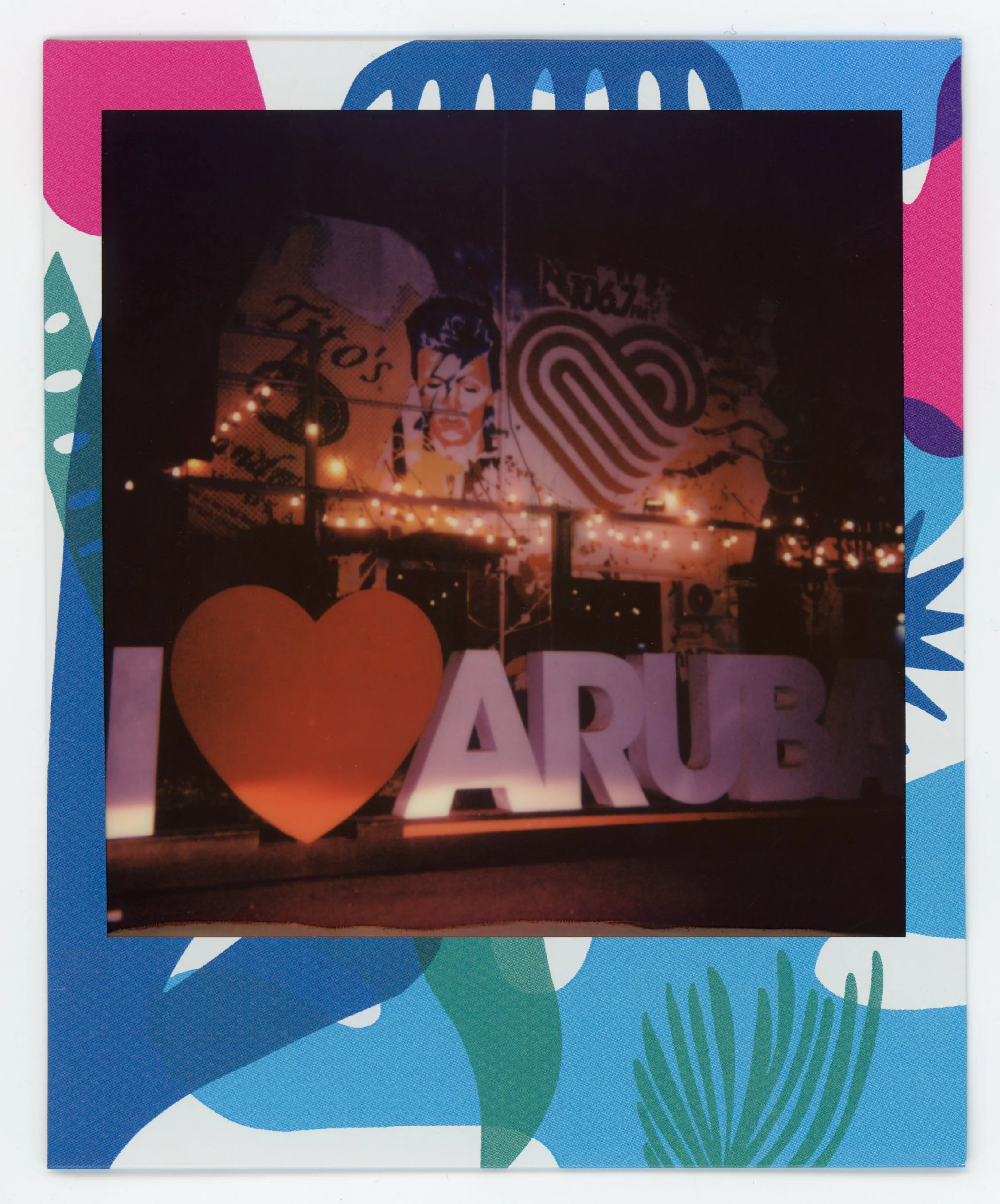 Photo of a sign that says 'I love Arub' with a heart symbol in place of the word 'love'. The background features a mural with a portrait and a heart-shaped design, along with string lights.