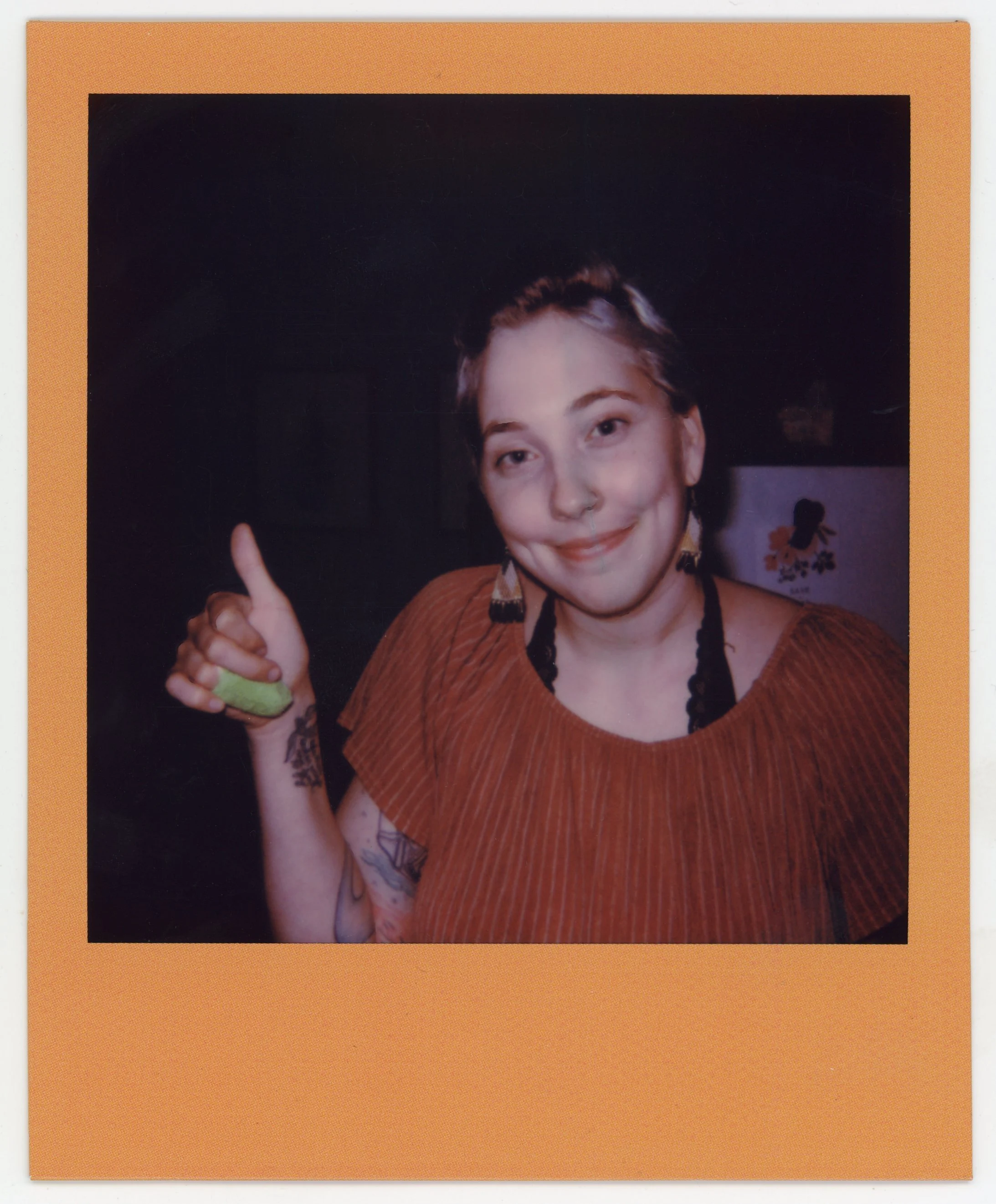 Young woman smiling and giving a thumbs-up, wearing an orange top and earrings, with tattoos on her arm and nose ring.