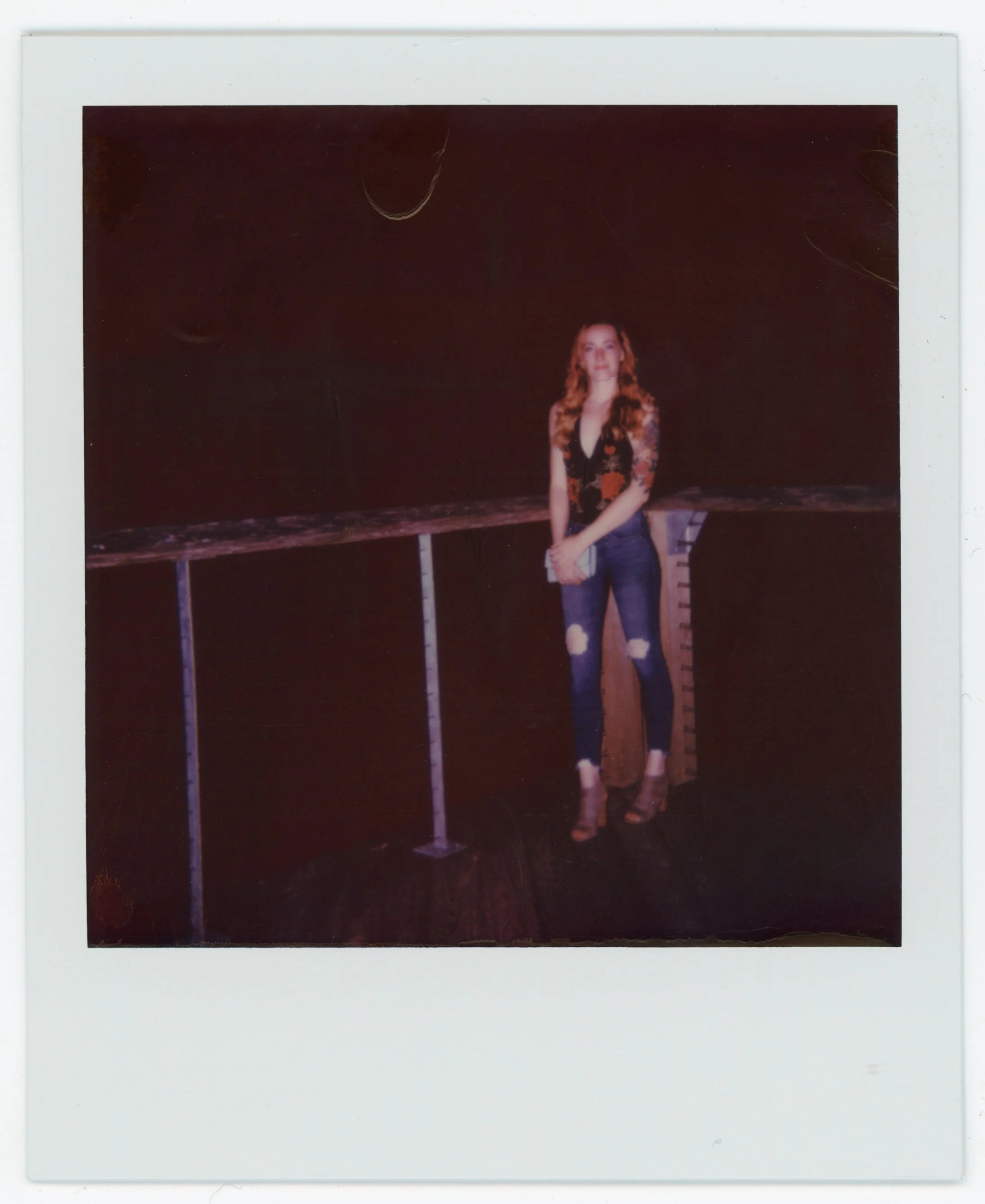 A woman with long, wavy hair standing in front of a dark background, wearing a black floral top, ripped jeans, and high heels, holding a small white item in her hands.