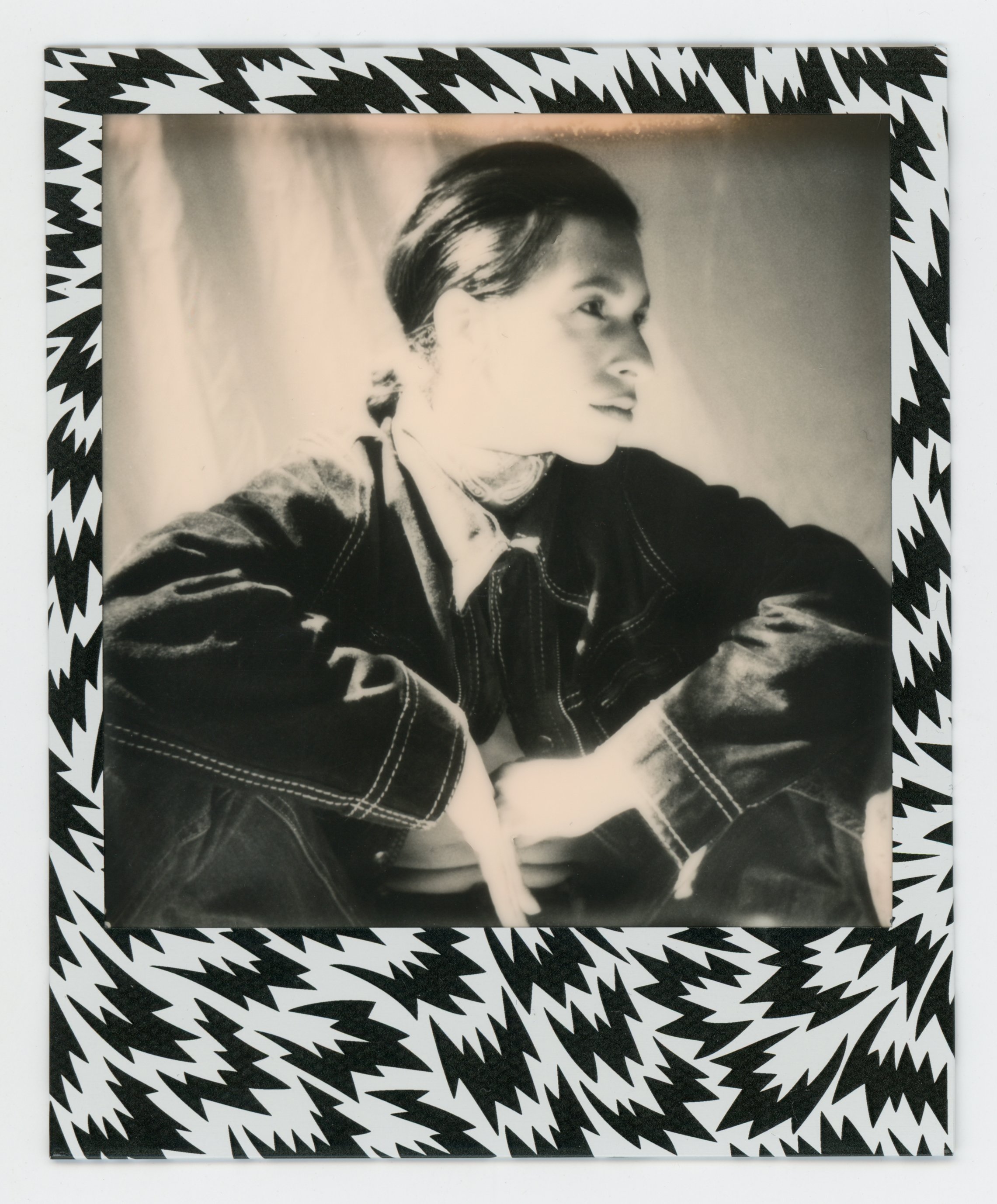 Black and white portrait of a woman with short hair, sitting with her arms crossed, framed by a zigzag patterned border.