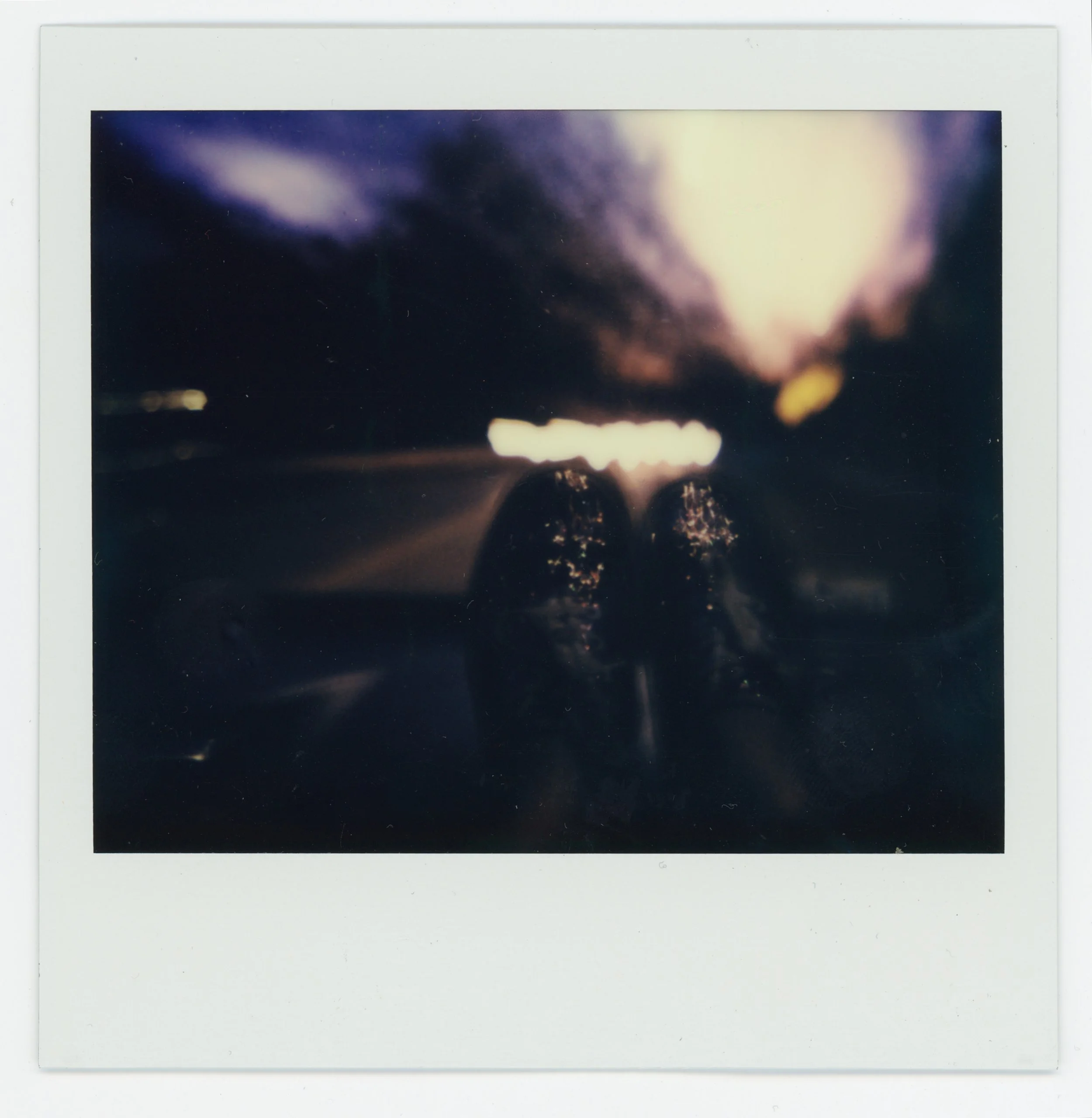A Polaroid photo of a person wearing dark sunglasses at night, with a dark sky and bright city lights in the background.