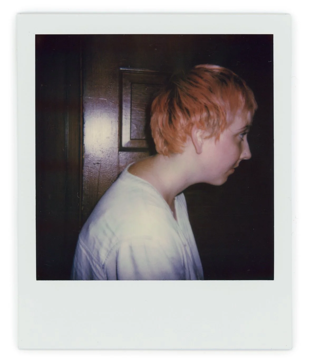 Side profile of a person with short red hair, wearing a white shirt, standing against a dark wooden wall.