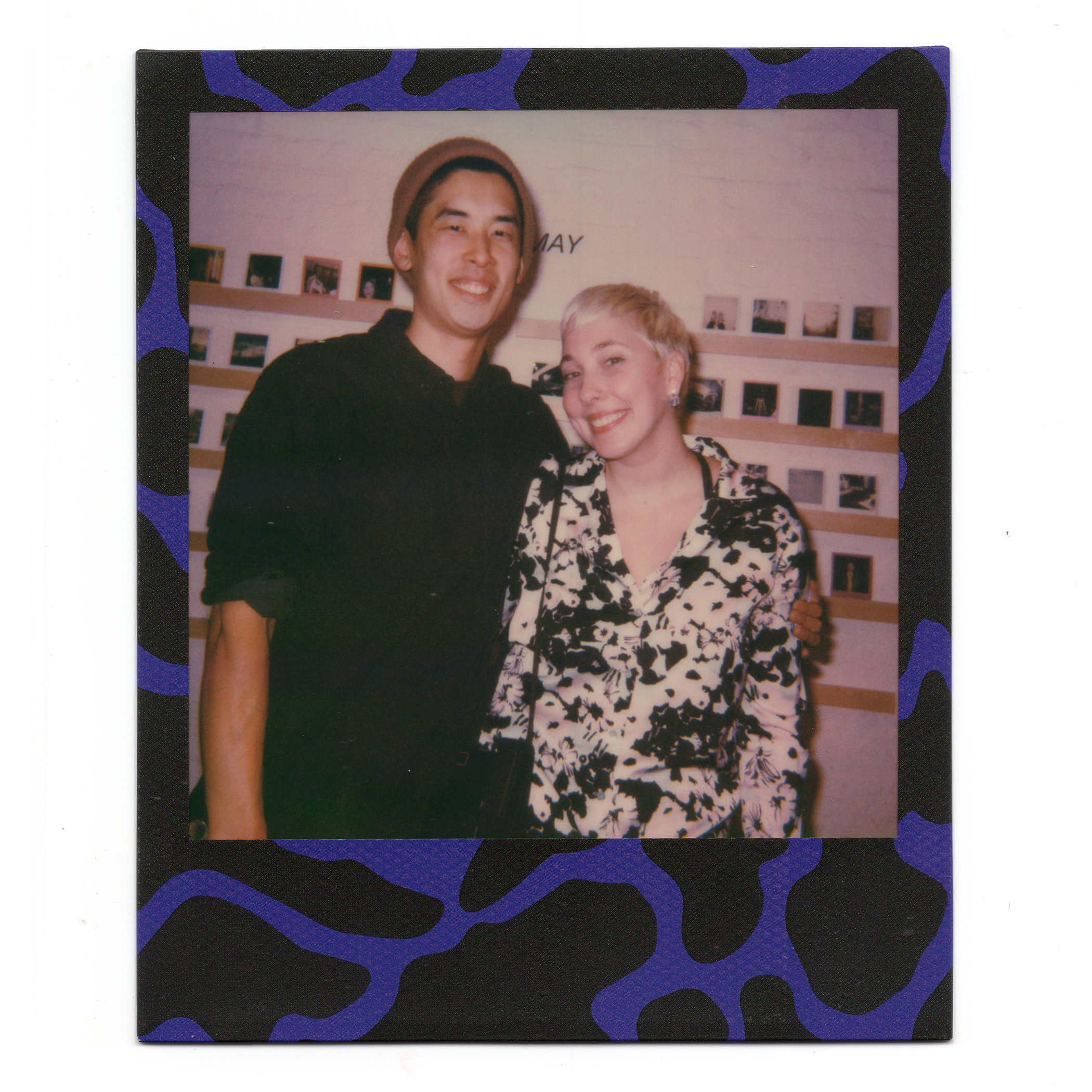 A young man and woman smiling and posing together inside a room with a wall display of small framed photos.