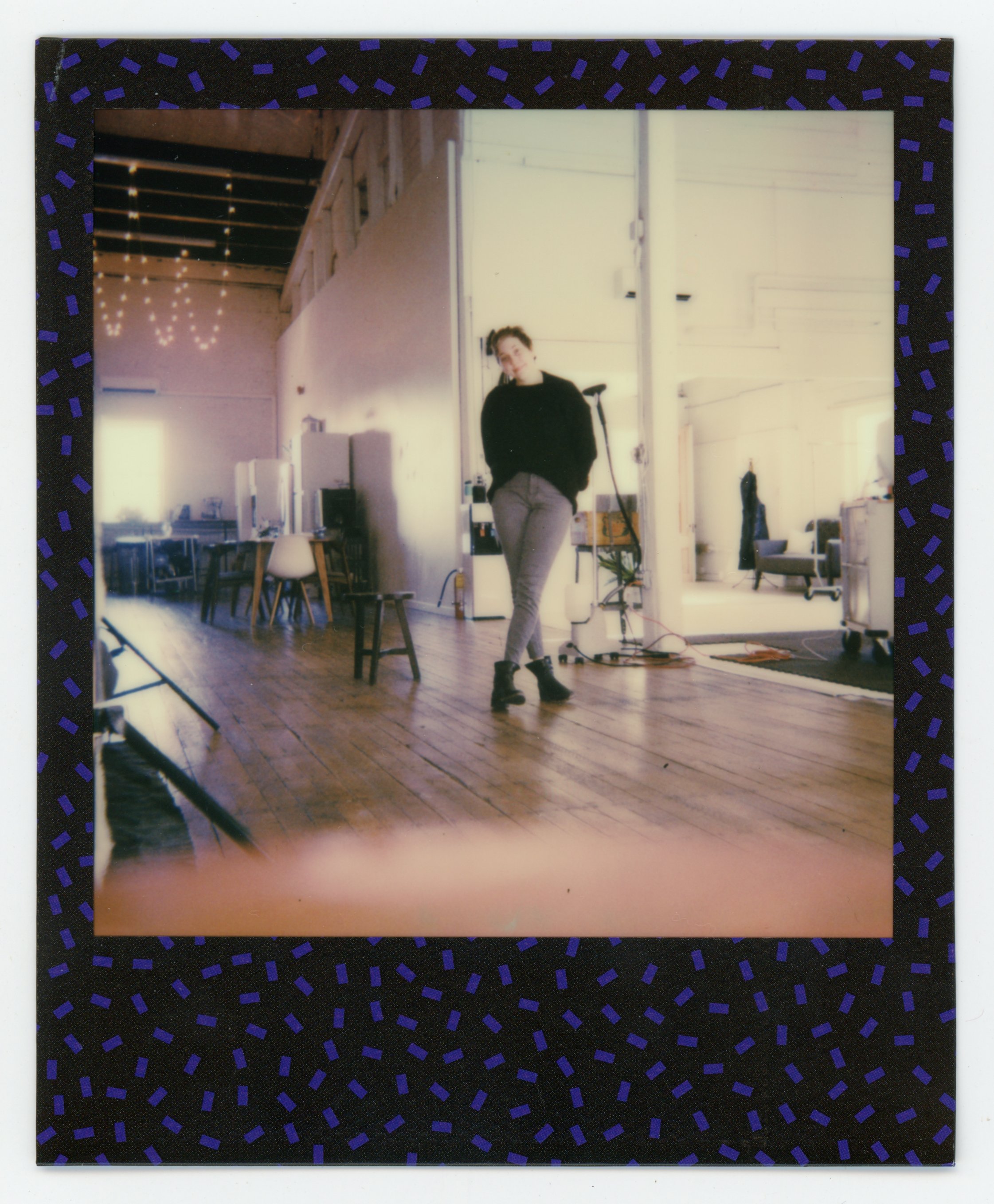 A woman standing in a spacious, well-lit room with wooden floors, holes in the wall and exposed ceiling beams, decorated with string lights and various furniture including chairs, a desk, and a sofa.