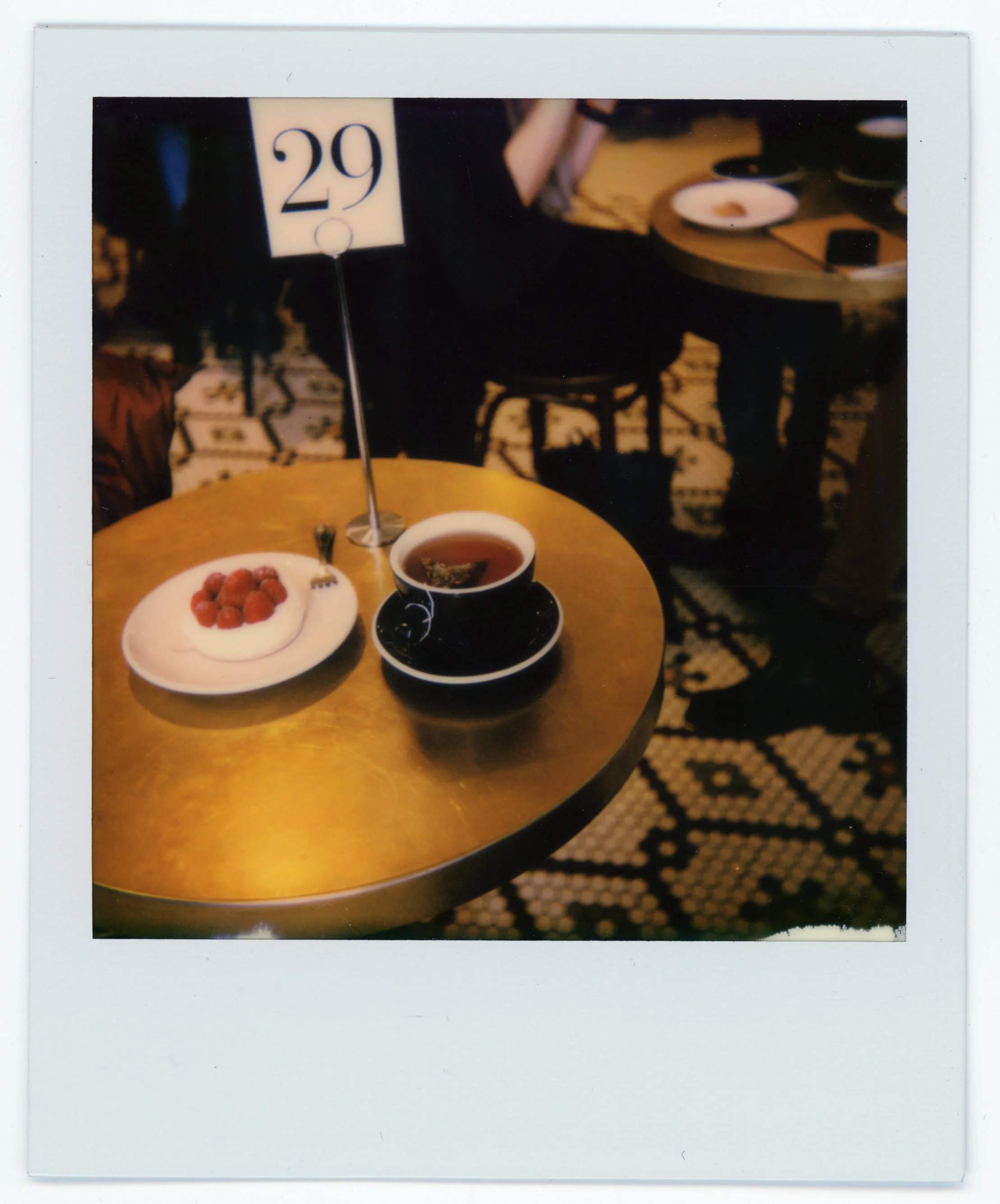 A round gold-colored table with a black cup of tea, a small plate of raspberries, and a silver fork. There is a table number sign with the number 29. In the background, other tables with similar settings are visible in a dimly lit room with patterned