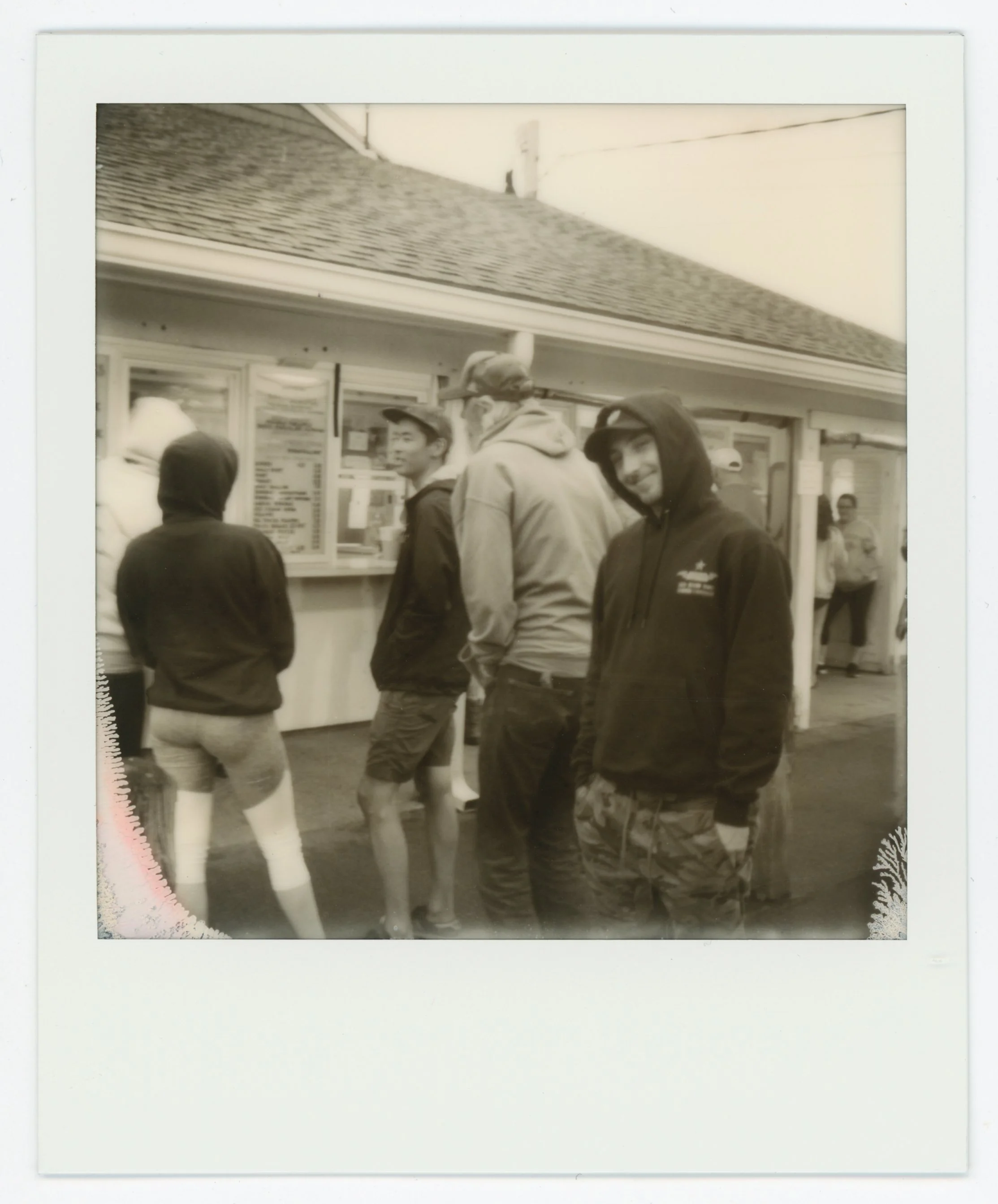 Group of young men in line at a fast food stand, wearing hoodies and shorts, some with hood up and some with face masks, in an outdoor setting.