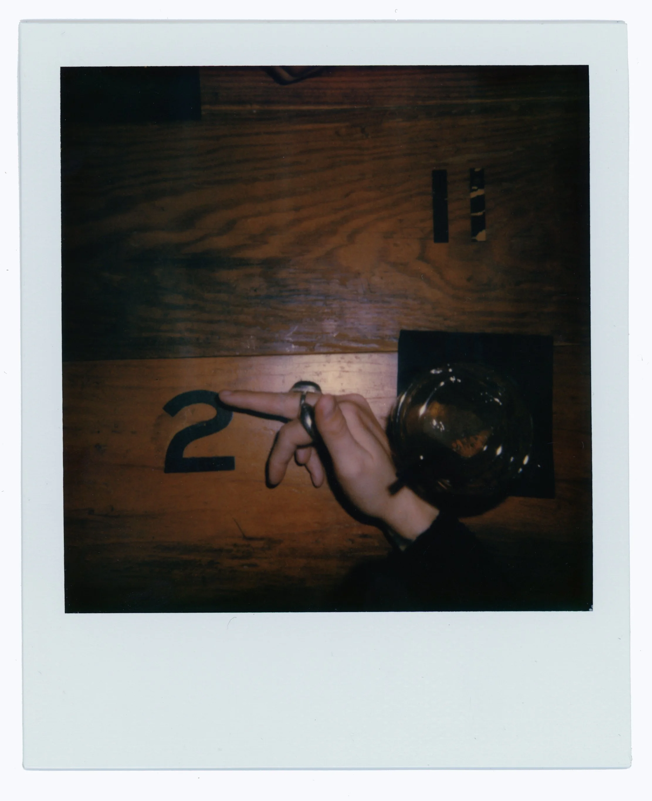 A hand with rings on fingers pointing to the number 2 on a wooden table, with a glass of dark-colored beverage placed on a black coaster next to it.
