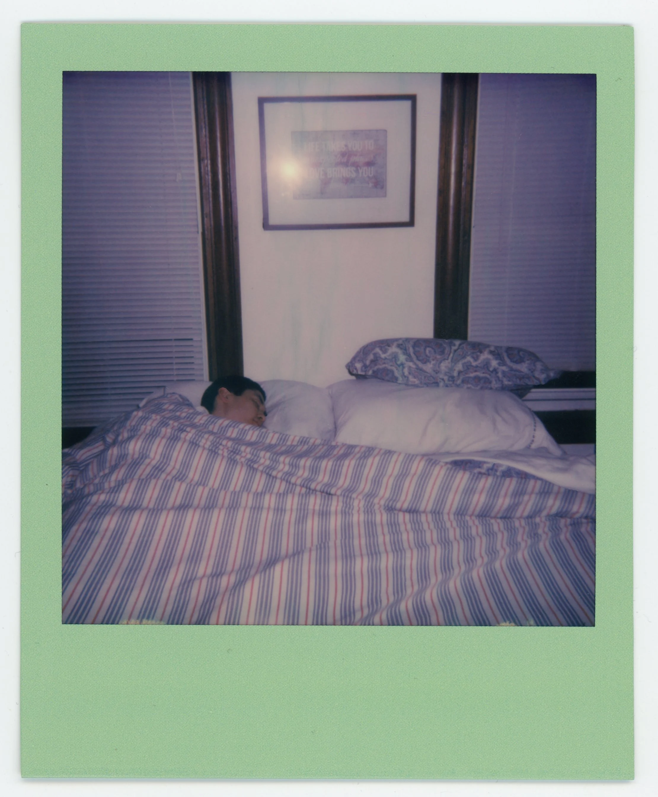 Person sleeping in bed on striped blanket with pillows, framed picture on wall in background.