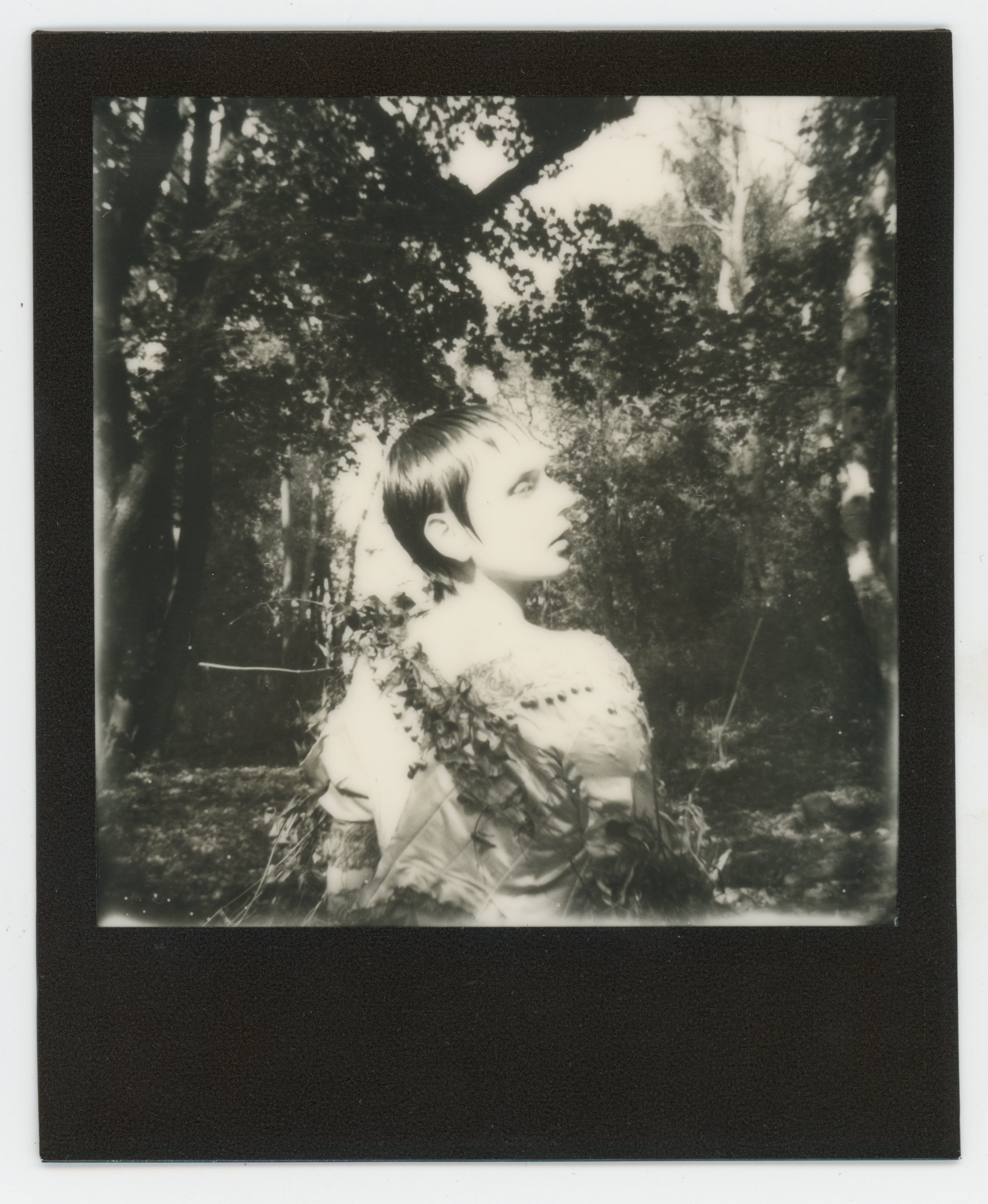 Black and white photograph of a woman with short hair, standing outdoors among trees and foliage, looking to her right.