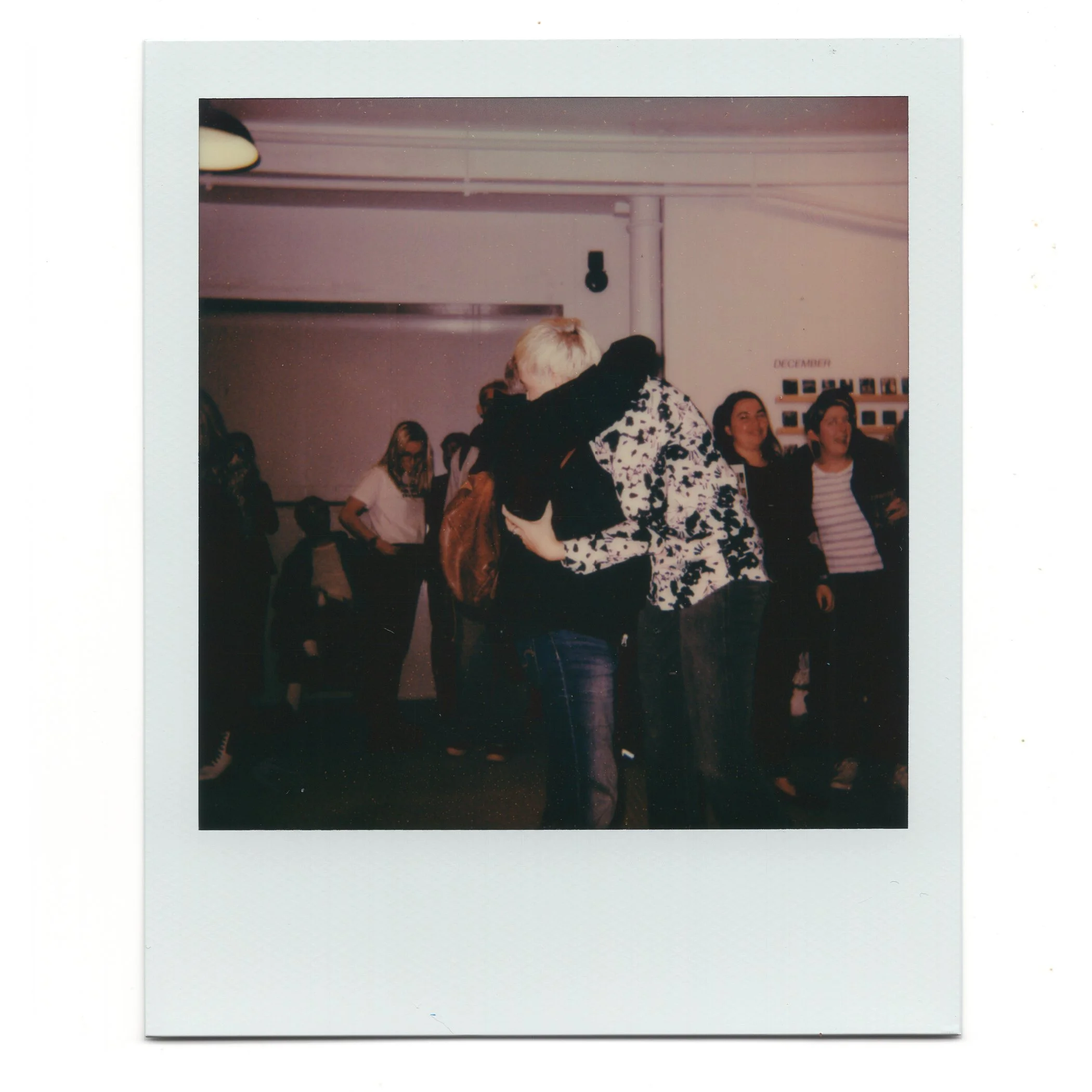 Two people hugging each other, surrounded by a group of people in an indoor setting, possibly a gallery or event space.