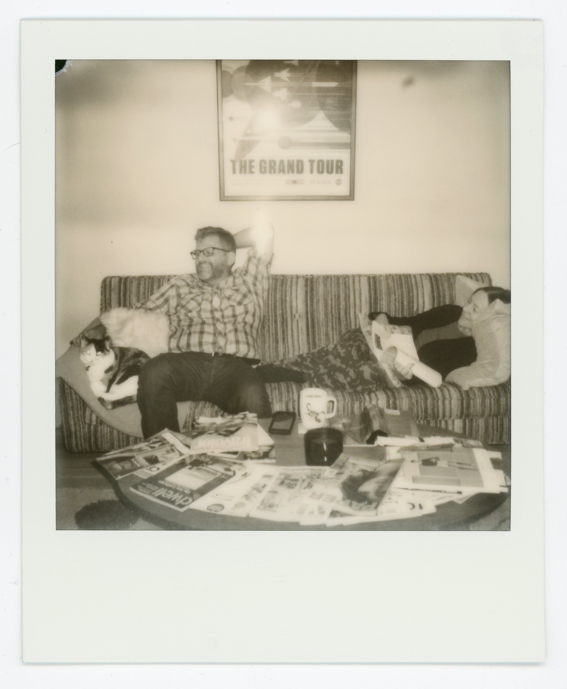 Two people relaxing on a striped couch; one man with glasses and a checkered shirt, and a woman reading a book; a cat sprawled on the man's lap; a coffee table in front with magazines, newspapers, a mug, and electronic devices; a framed poster on the