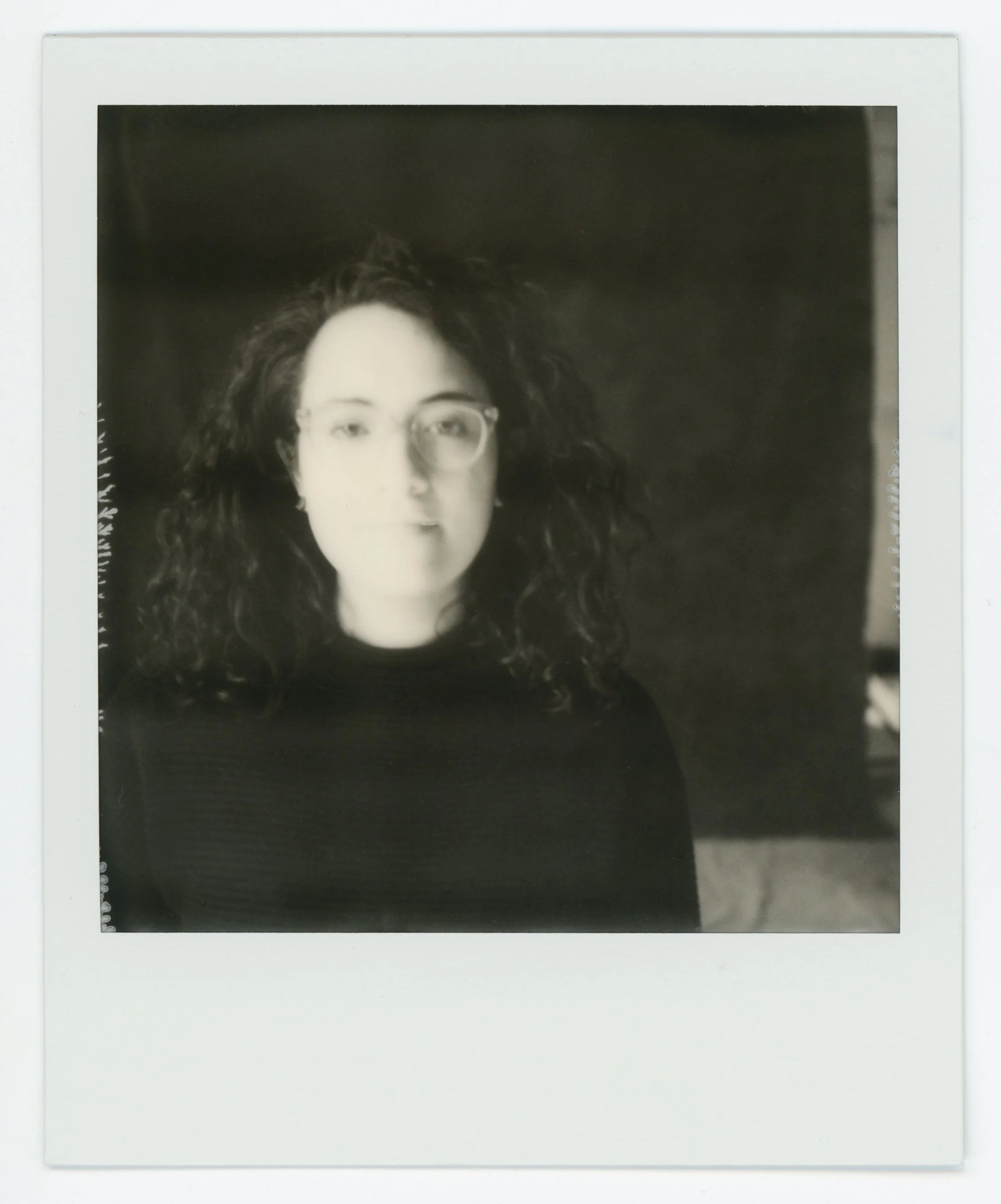 Black and white instant photograph of a woman with curly hair and glasses, wearing a dark top, against a dark background.