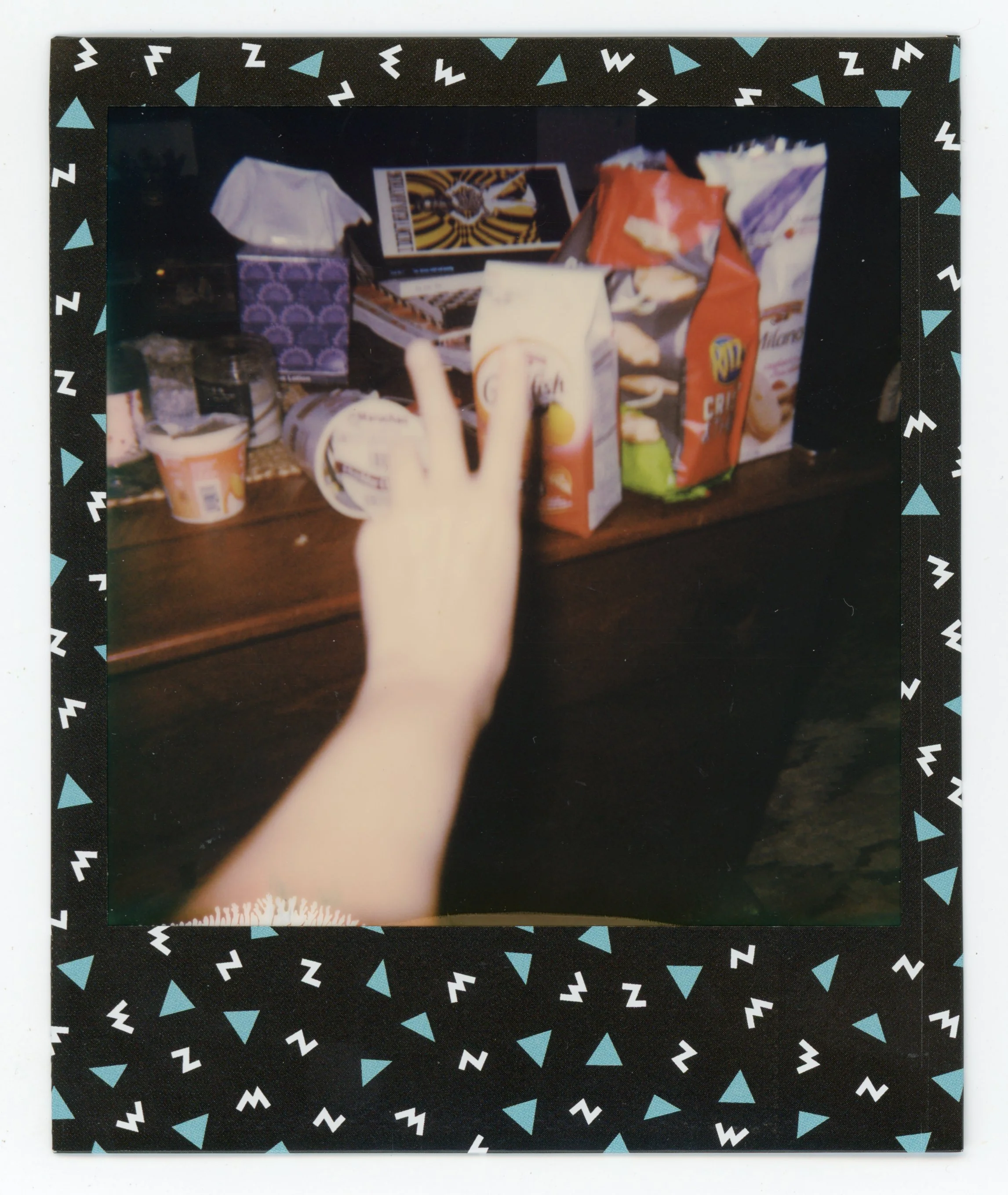A hand making a peace sign in front of a table with snack and drink items, including chips, popcorn, and juice boxes, in a room with a decorative border.