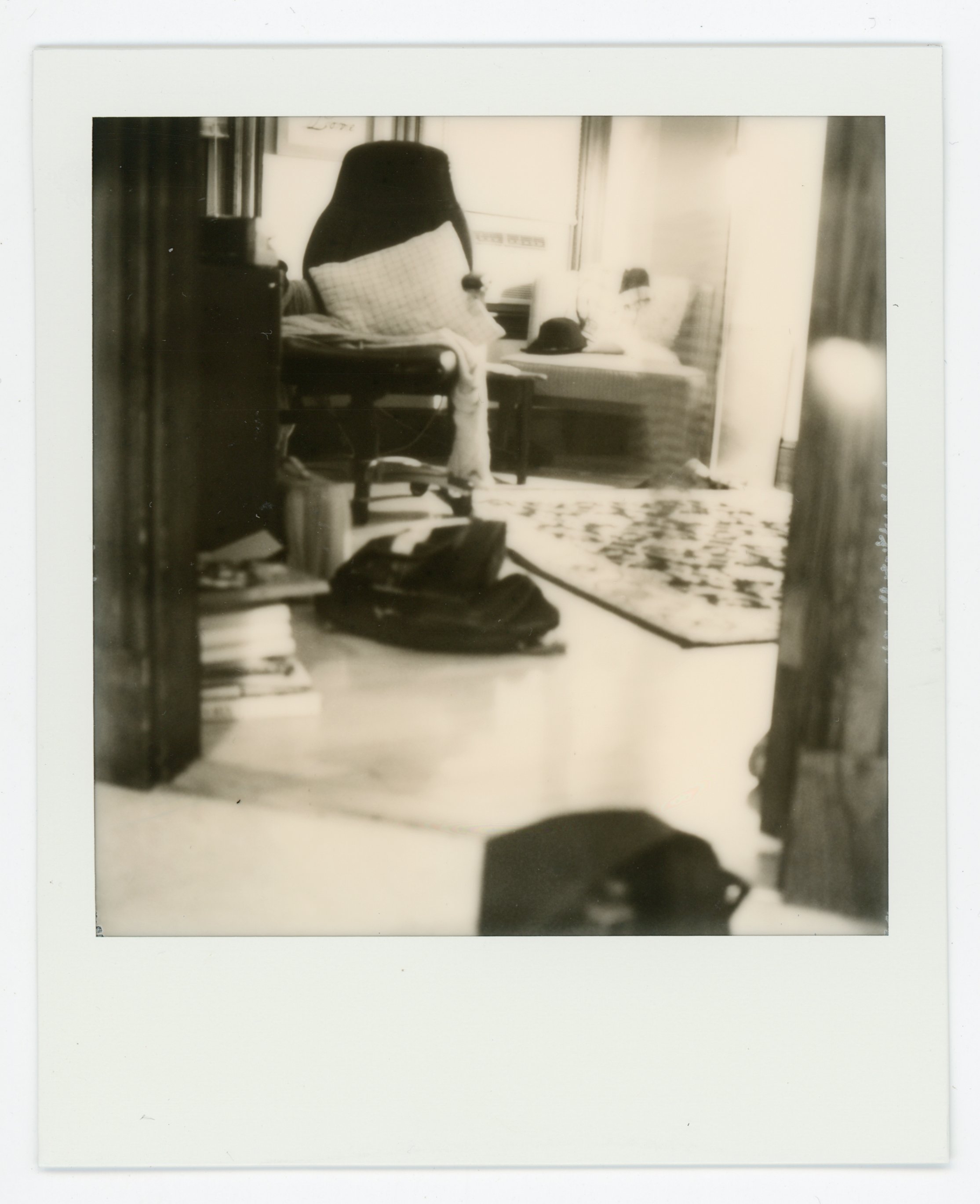 A black and white photograph of a cozy living space with a armchair, a bench with pillows, a rug, and various items like a hat and a bag, viewed from a doorway.