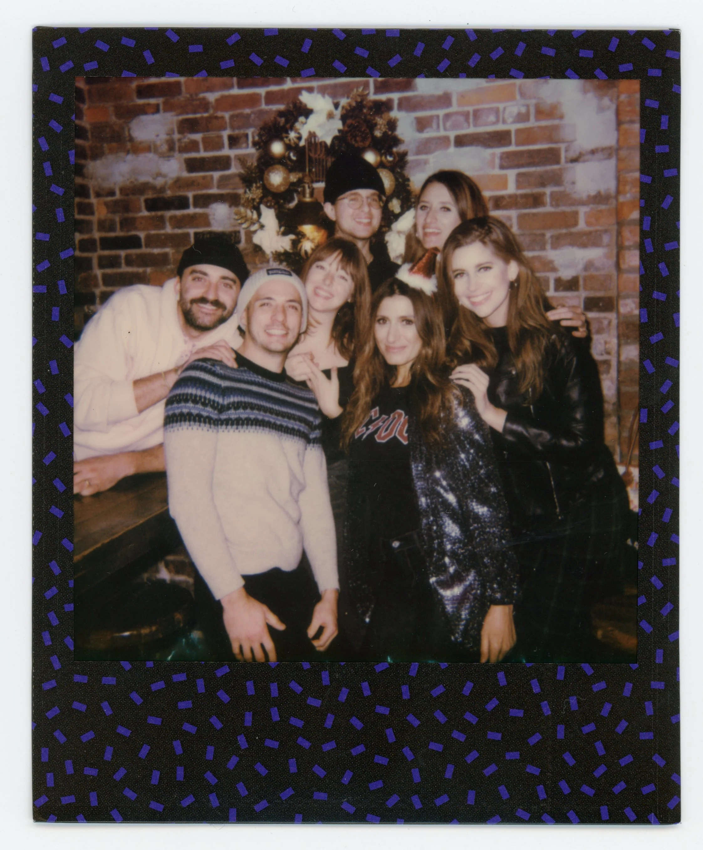 A group of seven friends gathered in front of a Christmas tree with ornaments and flowers, smiling for a photo at a holiday celebration.
