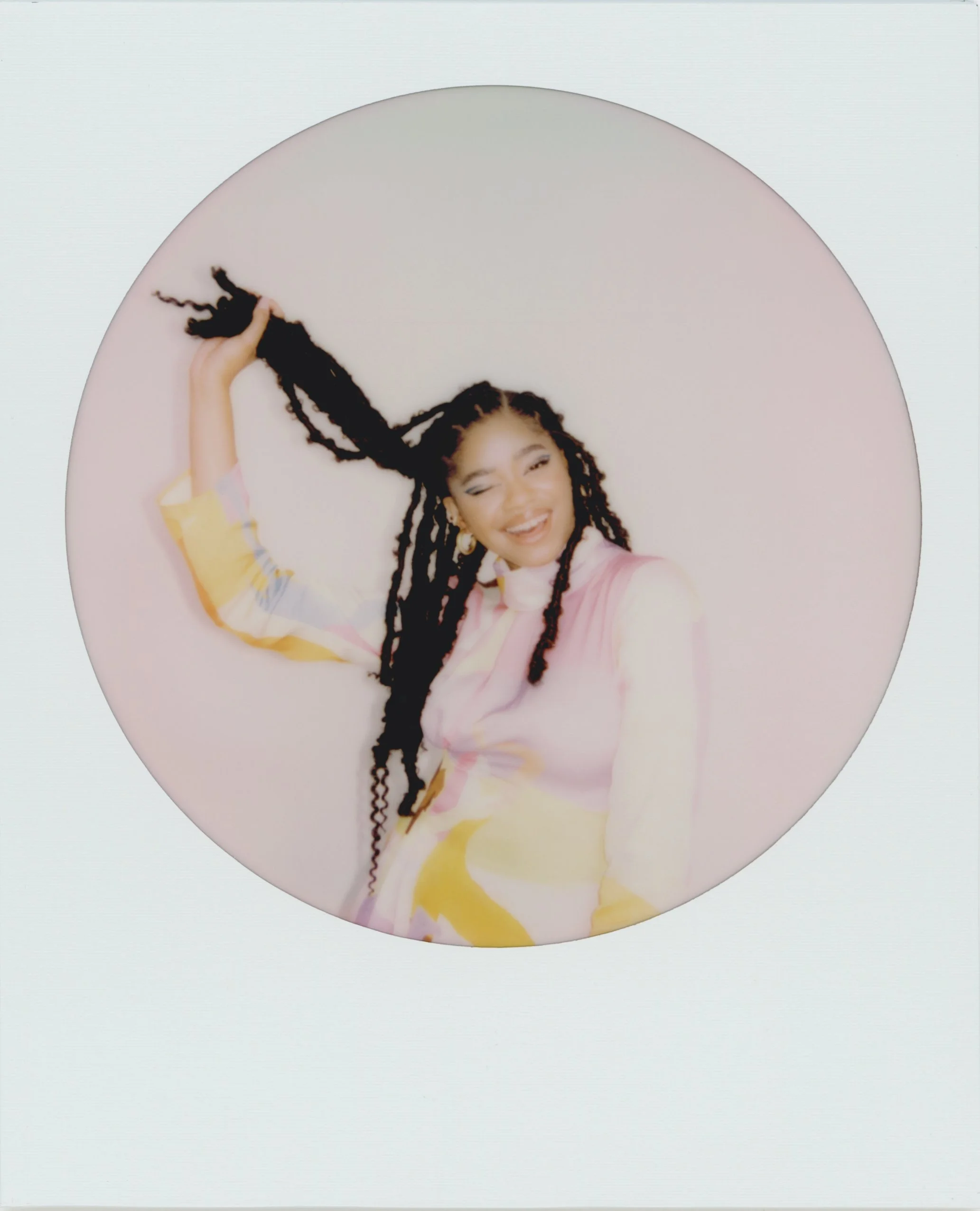 A woman with long, braided hair poses against a light pink background, wearing a pastel-colored dress and smiling while holding a section of her braids.