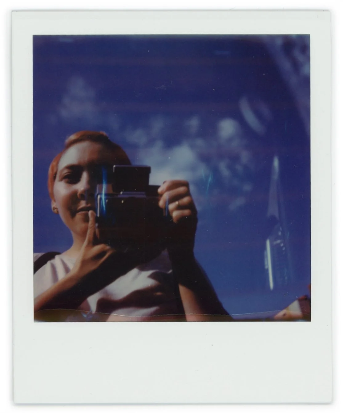 A woman taking a selfie outdoors on a sunny day, with blue sky and clouds reflected in a convex mirror or reflective surface.