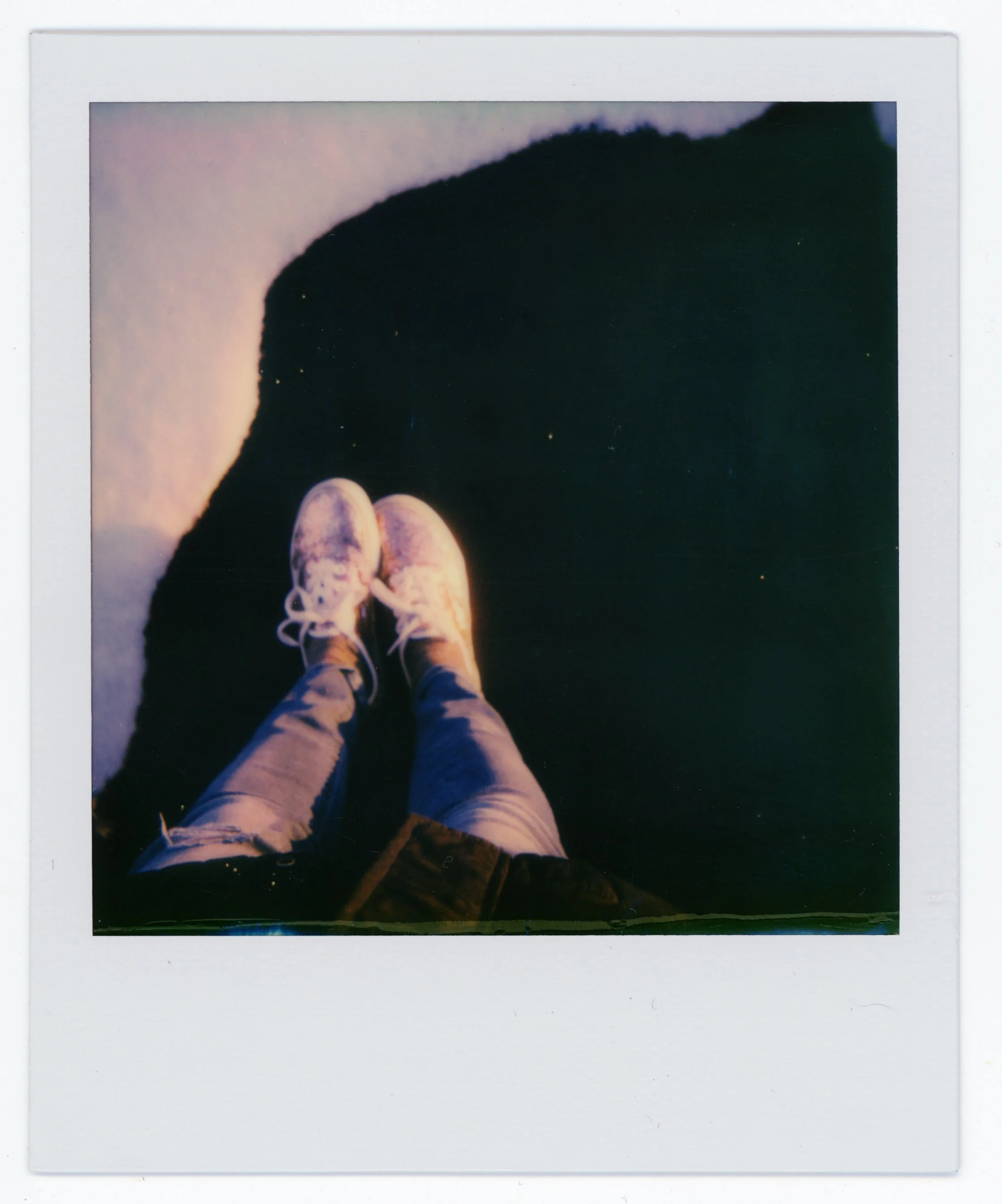 A person standing on a dark surface, wearing light-colored sneakers and jeans, looking down at their feet.