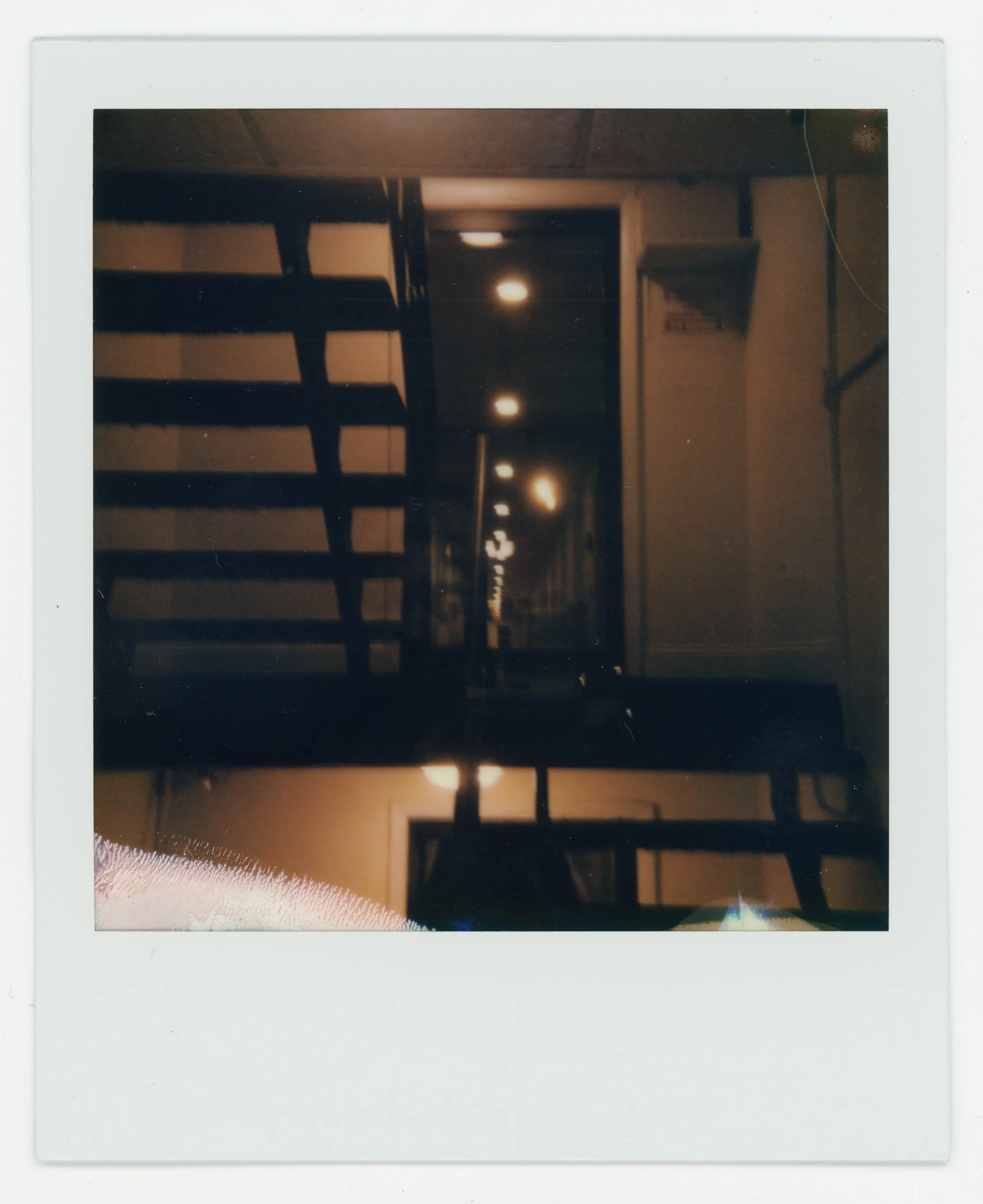 Dimly lit interior of a building with a staircase, with ceiling lights and a corridor visible in the distance.