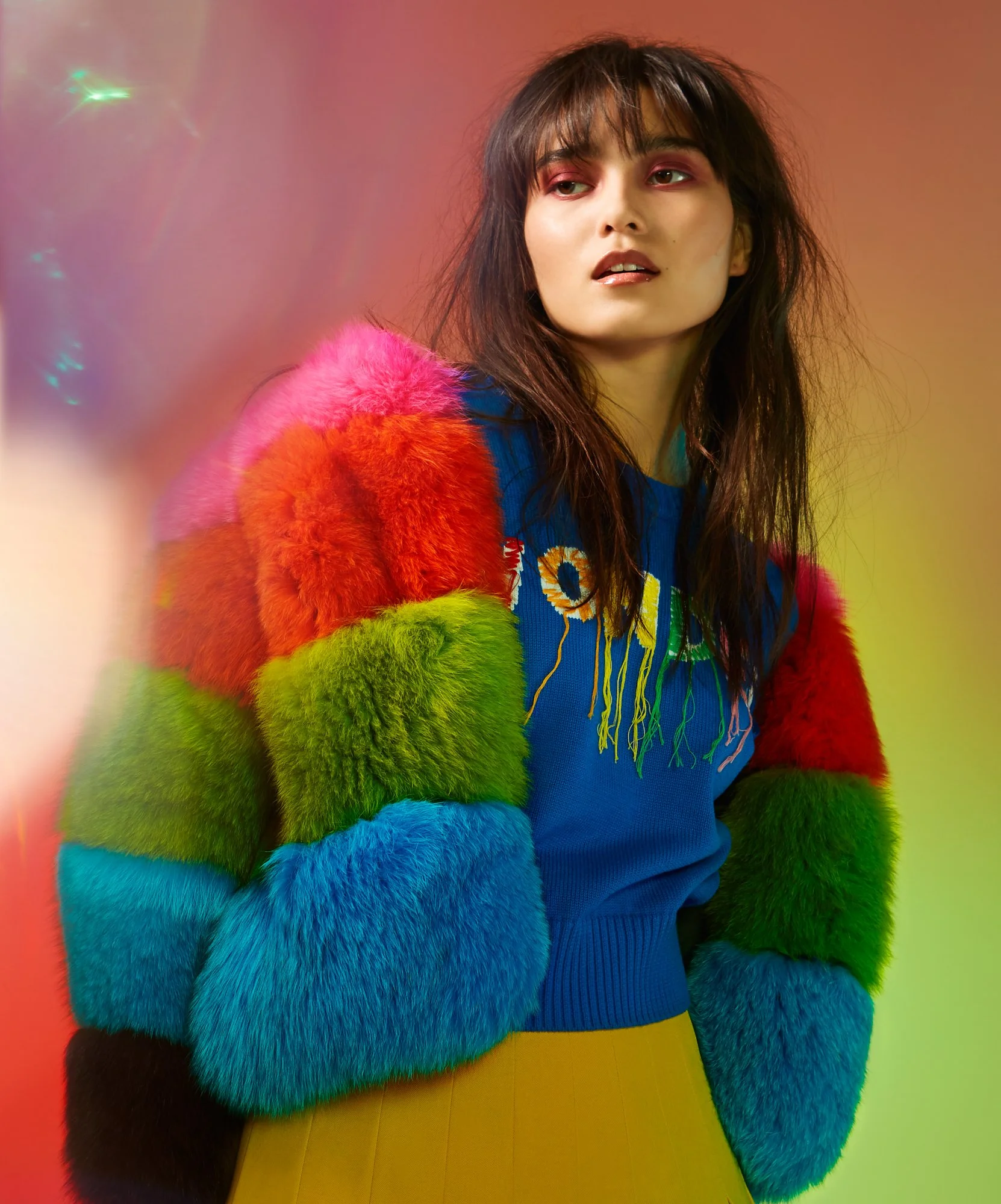 A young woman with dark brown hair wearing a vibrant rainbow-colored faux fur jacket and a blue embroidered sweater against a colorful, blurred background.