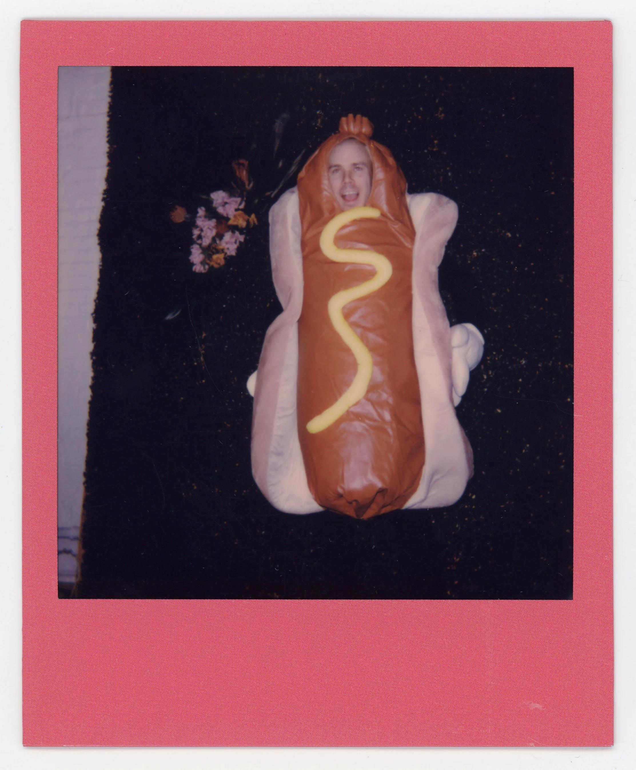 A person lying on a black surface wearing a hot dog costume with a yellow mustard squiggle, surrounded by some flowers and a white wall.