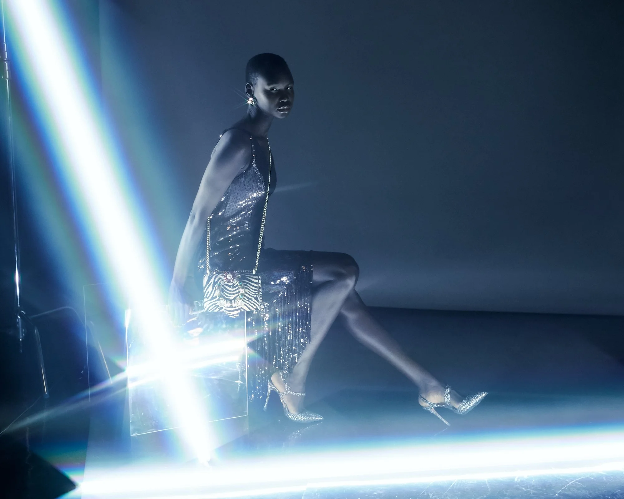 A woman with short hair wearing a shimmering dress and high heels, sitting on a reflective surface in a dark room illuminated by bright, colorful lights.