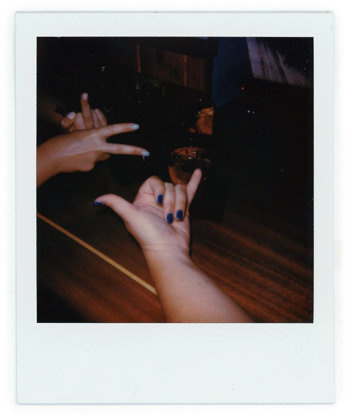 Two hands with painted nails making peace signs and one hand pointing at a glass on a wooden table, with a blurry background.