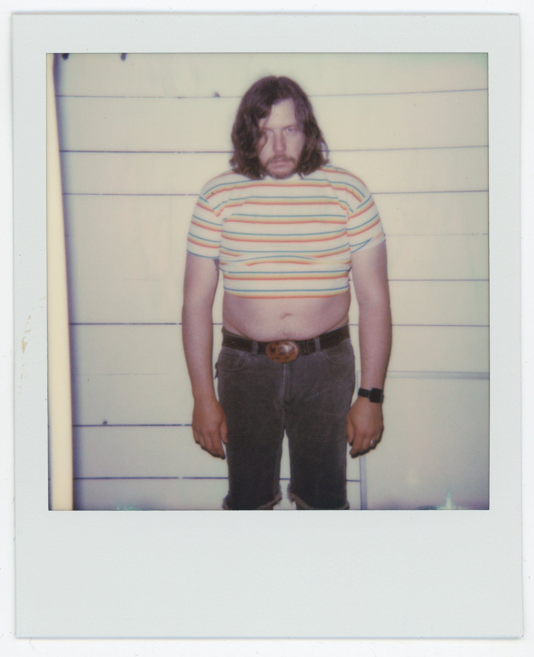 A man with long hair and a beard standing against a light-colored wall with horizontal paneling, wearing a striped crop top, dark pants, a belt, and a watch.