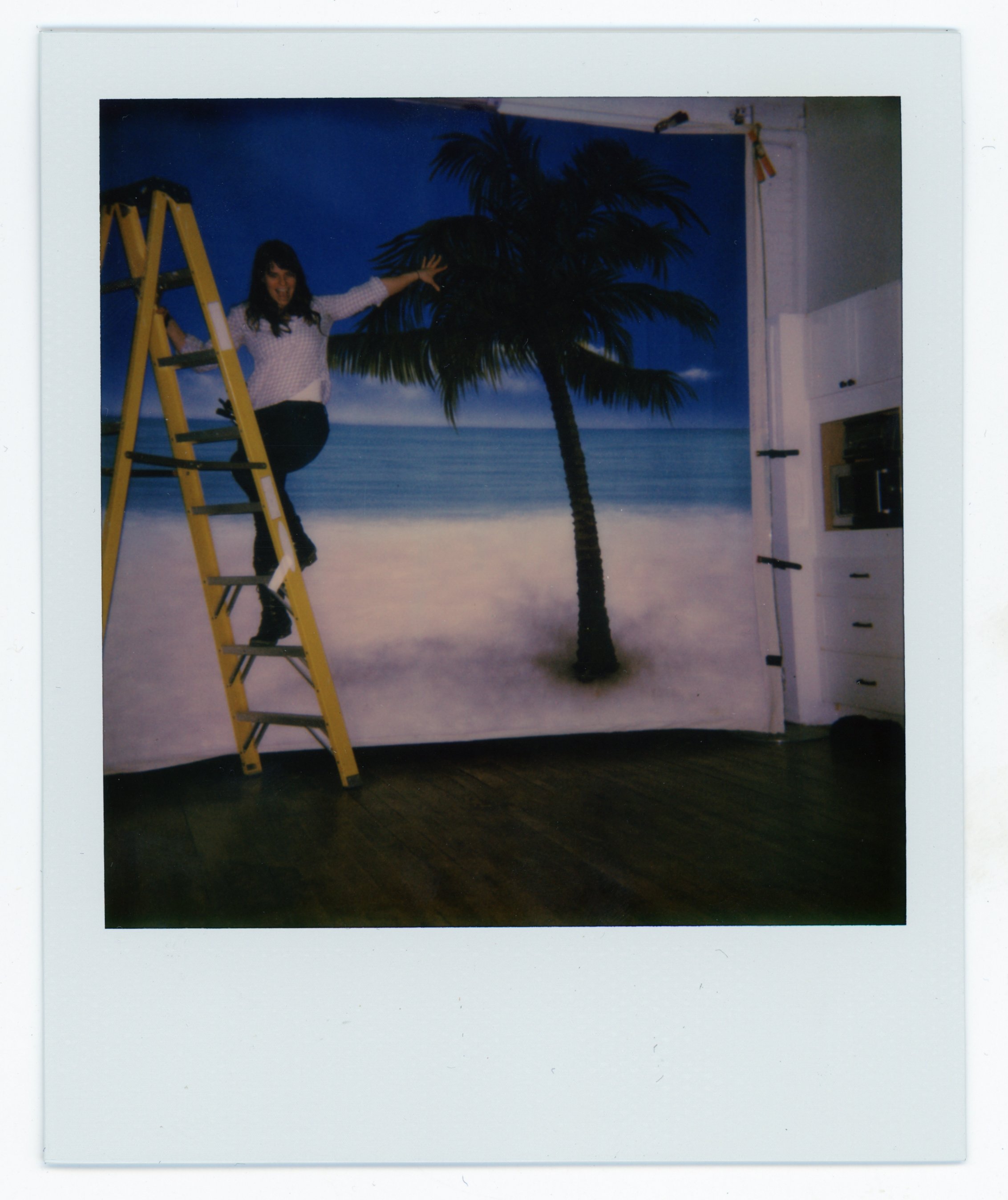A woman standing on a yellow ladder, pointing towards a beach scene backdrop with a palm tree, ocean, and sand, indoors.