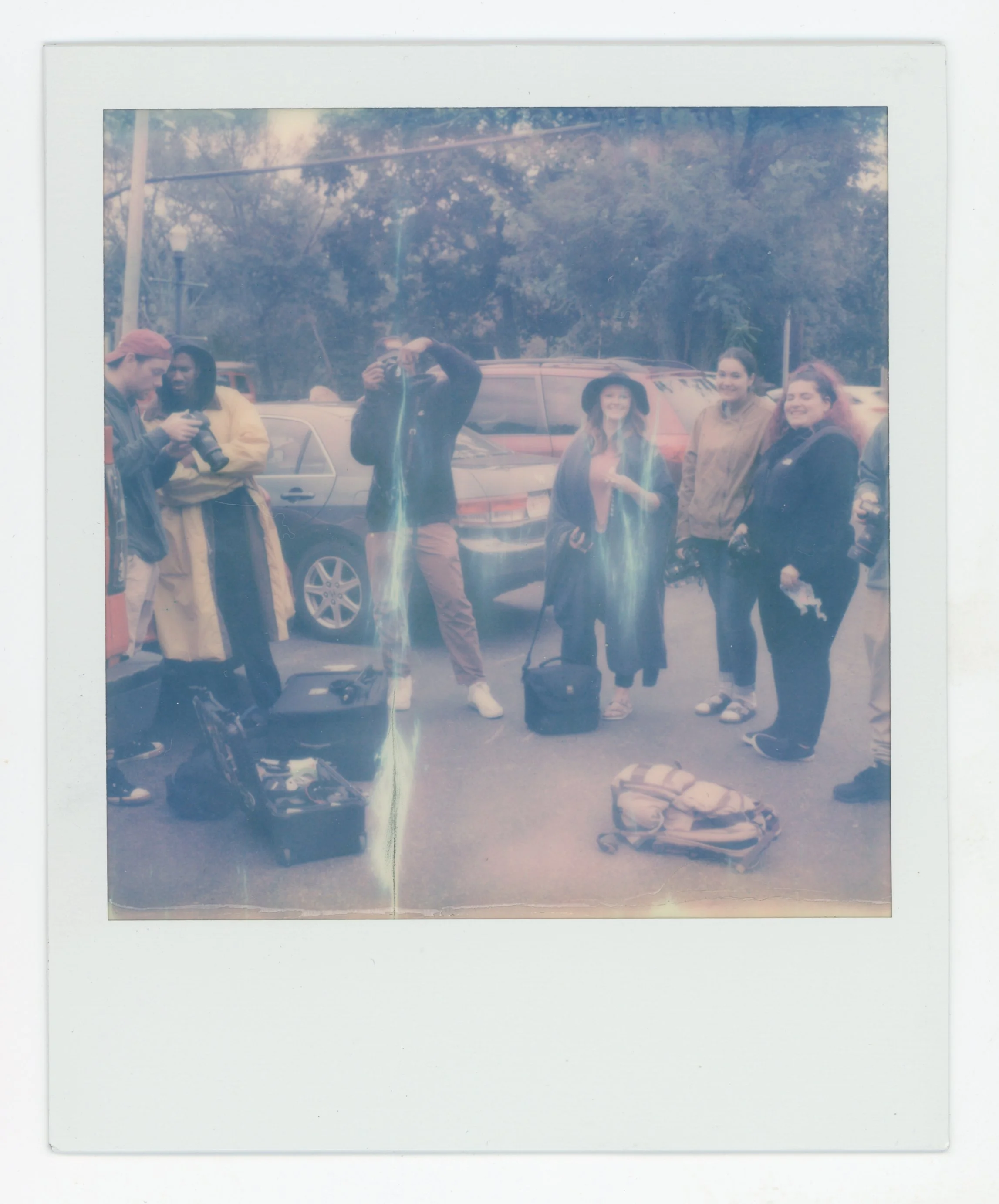 A group of six people standing outdoors in a parking lot, with cameras and equipment, some holding cameras, and two backpacks on the ground in front of them. The background includes trees and parked cars.