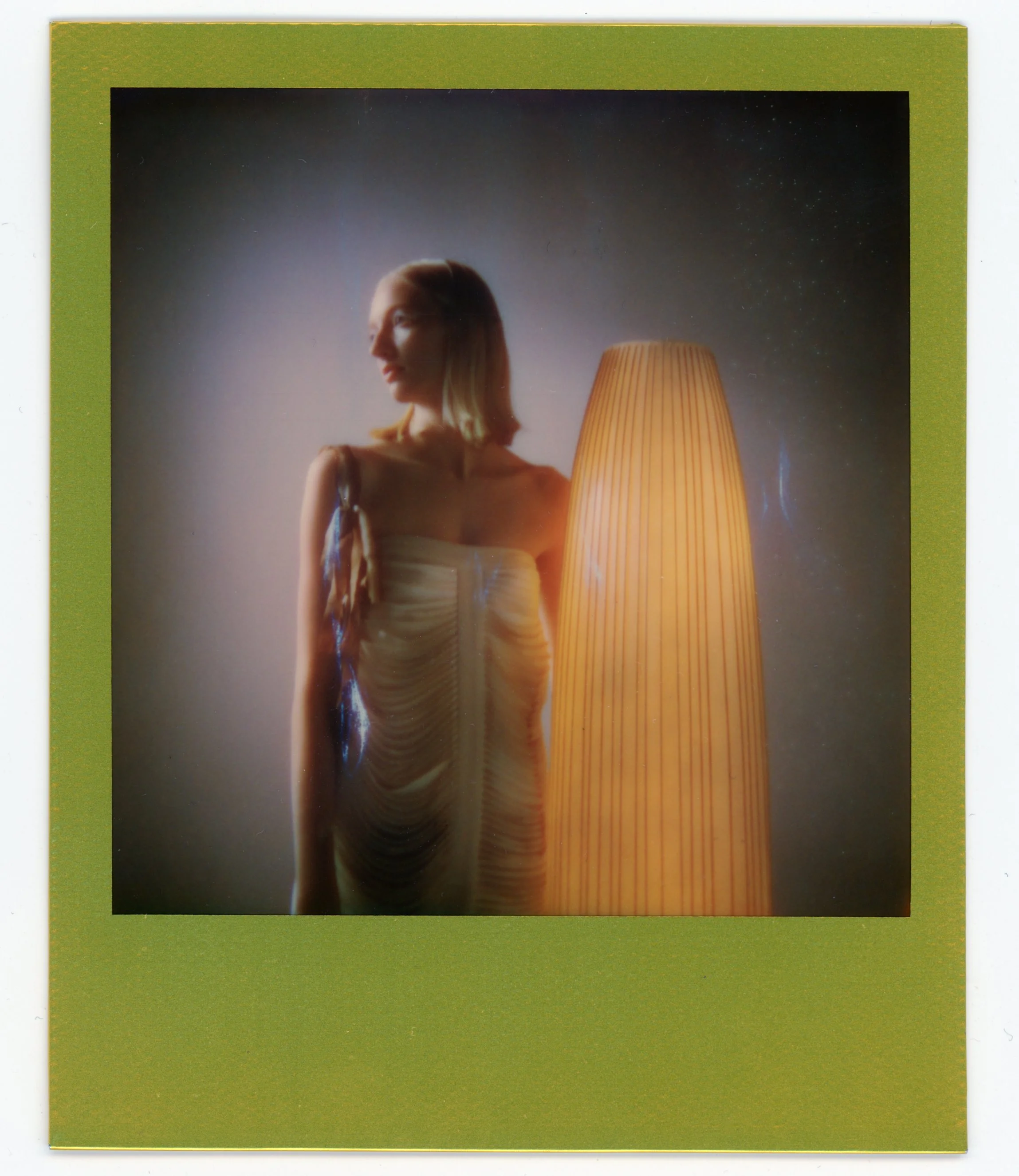 A woman wearing a strapless dress stands next to a tall, orange lampshade, with soft lighting creating a gentle glow and a blurred background.