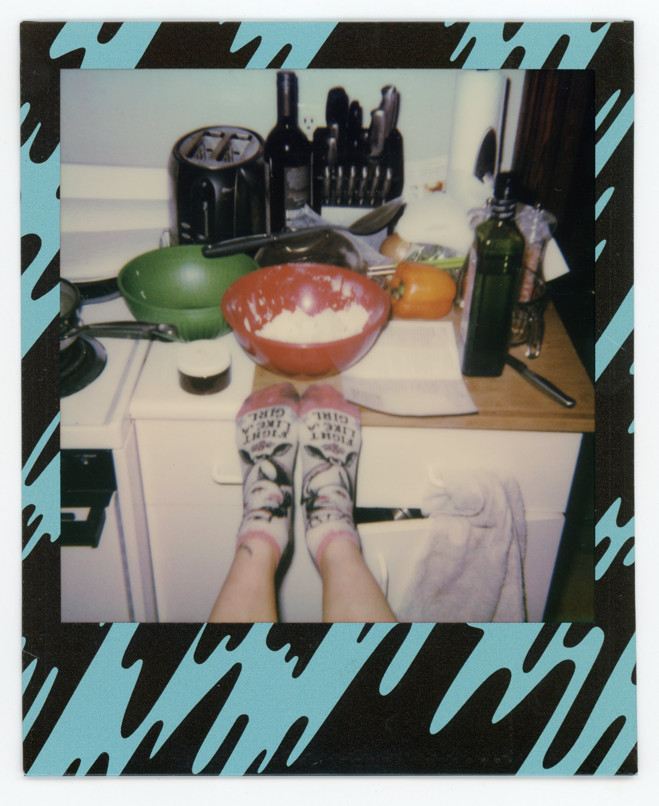 First-person view of a person's legs and feet wearing socks with text, standing in front of a kitchen counter with various kitchen items, including a red mixing bowl with dough, a green bowl, a yellow pepper, a bottle of olive oil, and other utensils