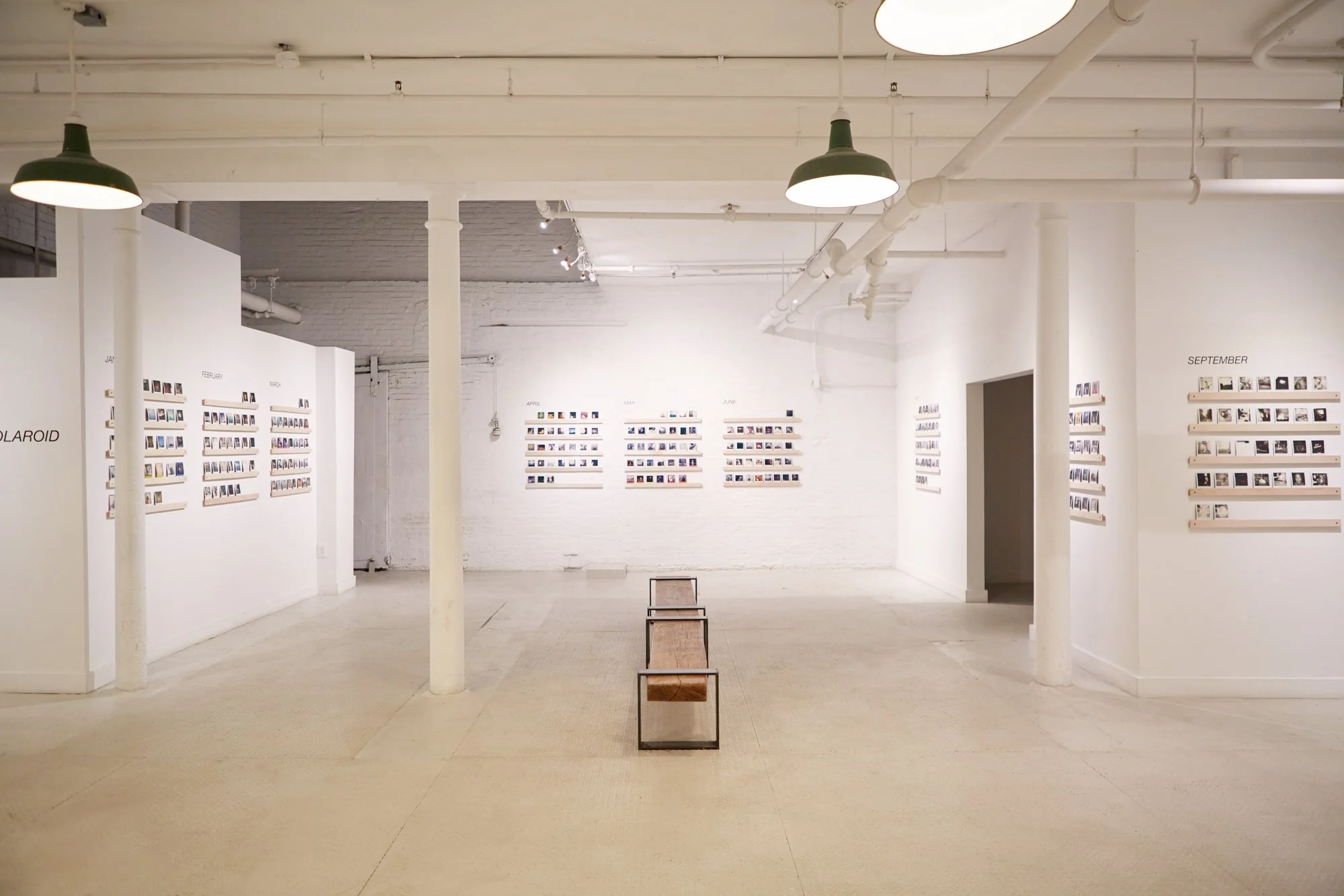 Empty art gallery with white walls displaying photographs organized by month, with a single wooden bench in the center and green pendant lights hanging from the ceiling.