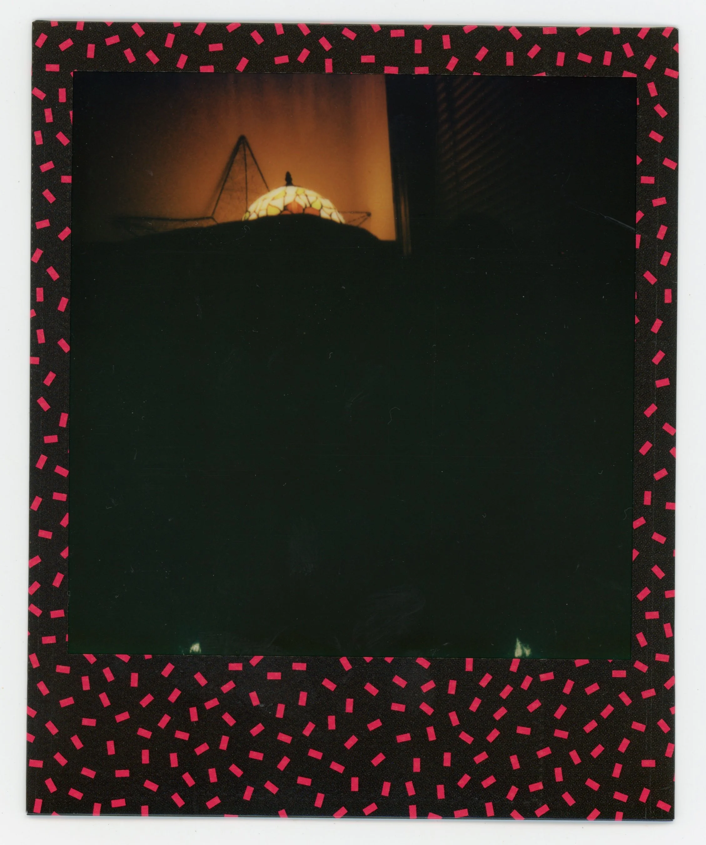 A dark square photo bordered by black with pink dashed lines pattern, with a lit lampshade visible at the top center, showing a warm glow and a small decorative object on top.
