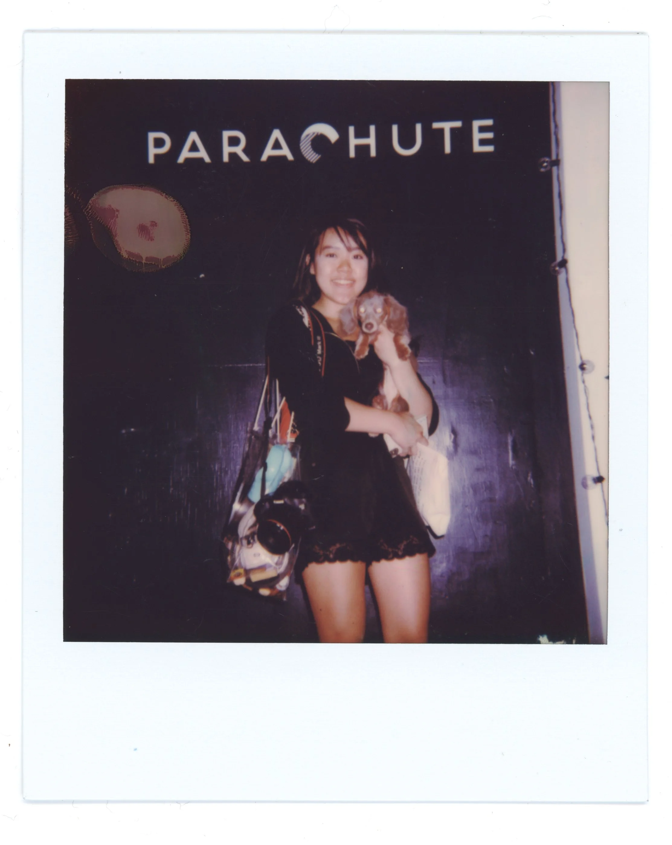 Young woman holding a small dog in front of a black wall with the word 'PARACHUTE' and a moon logo.
