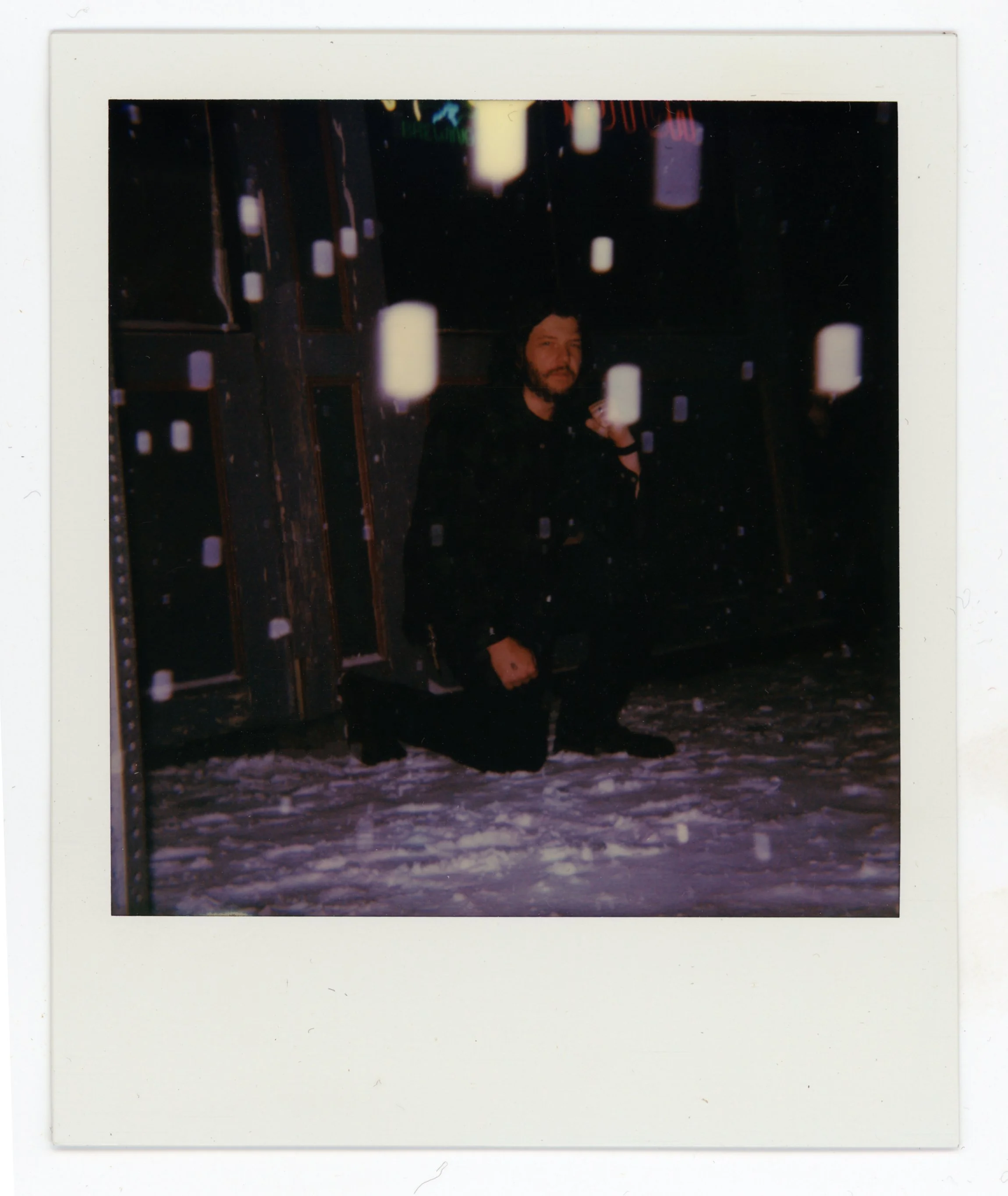 A person with a beard and dark hair squatting on snow with a drink in hand, in a dark room with wooden and metal walls, illuminated by scattered white light sources.
