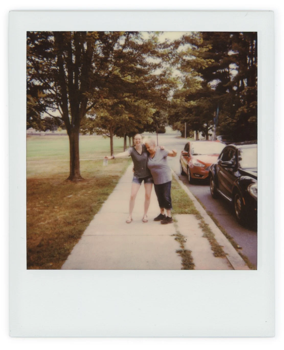 Two women standing on a sidewalk hugging, with parked cars and trees in the background.