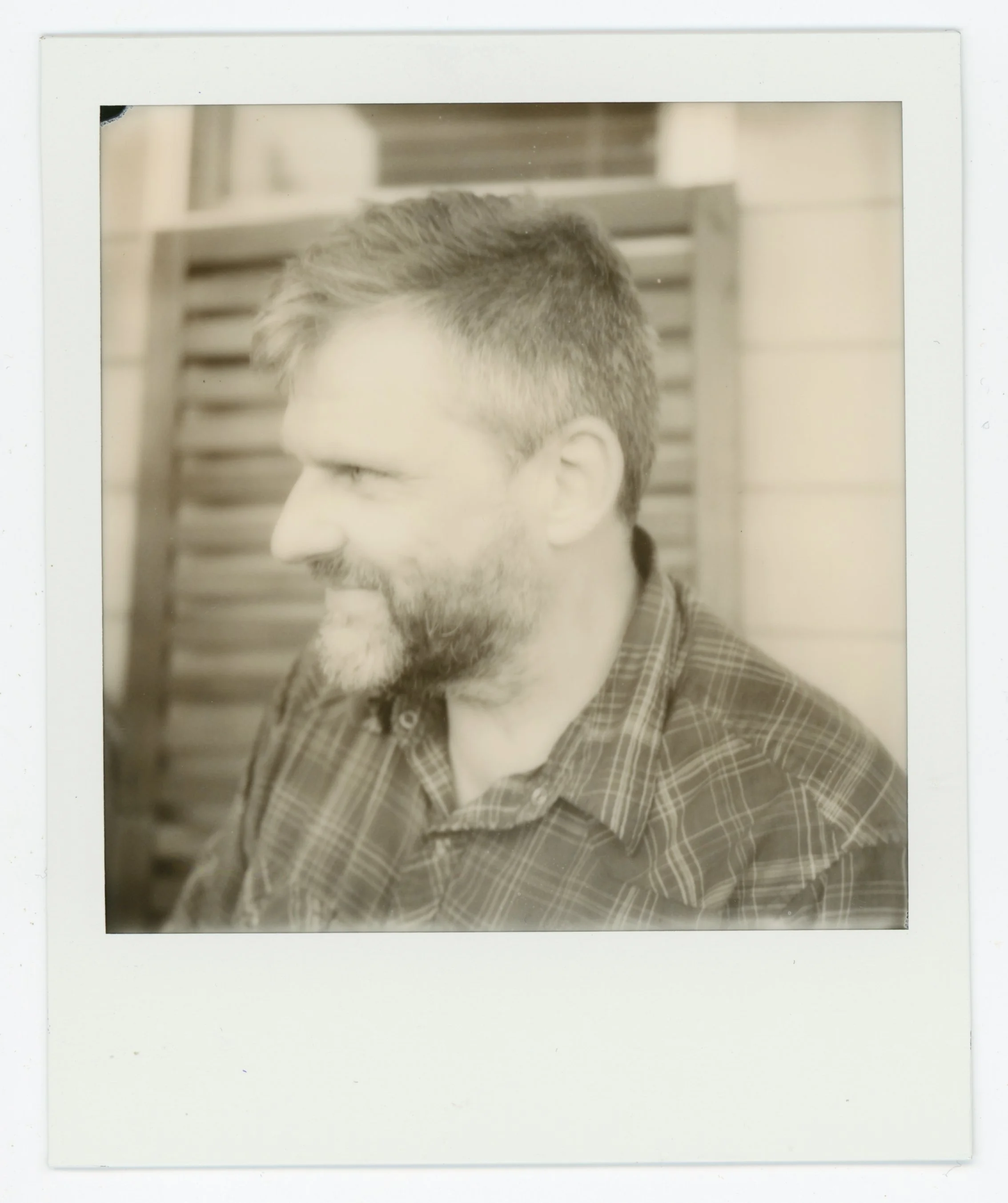 Black and white Polaroid photo of a man with a beard and short hair, wearing a plaid shirt, smiling and looking to the left, with a wooden fence in the background.