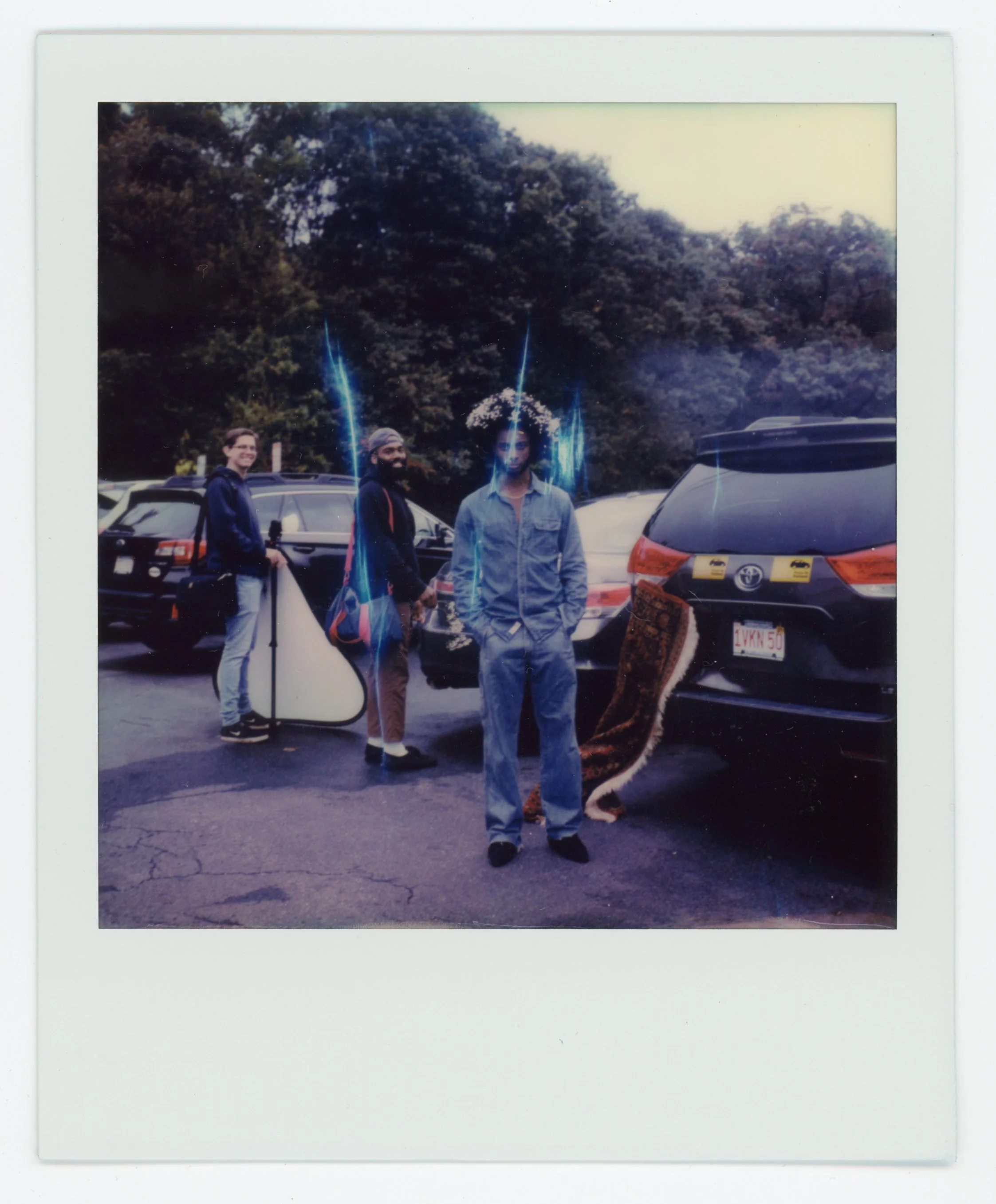 Three men standing in a parking lot among parked cars, one with a floral wreath on his head, and a fourth person holding a reflector. The photos have American car license plates, trees in the background, and blue light streaks overlaying the image.