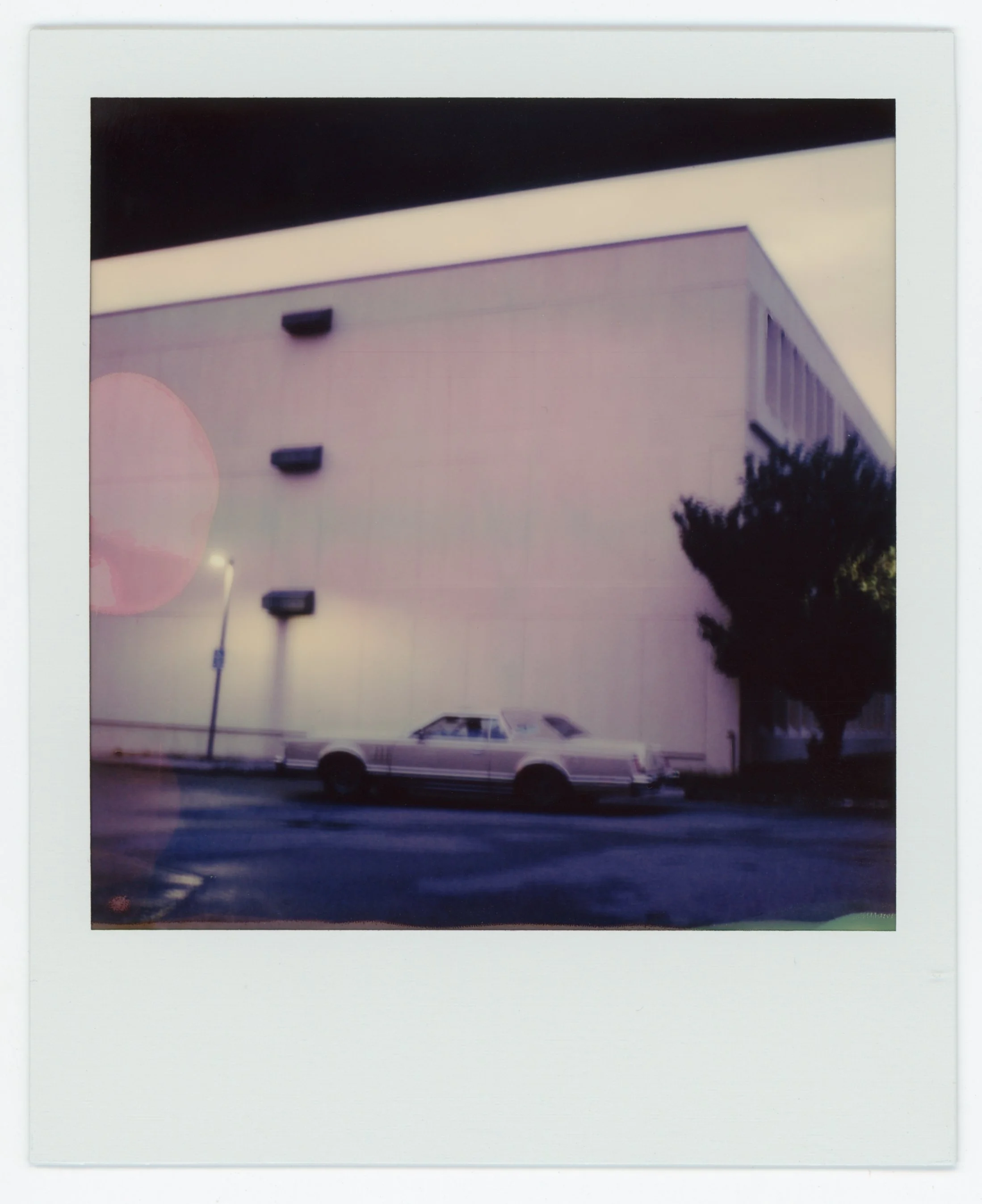 A vintage car parked outside a large beige building with air conditioning units and trees, under a black and white sky.