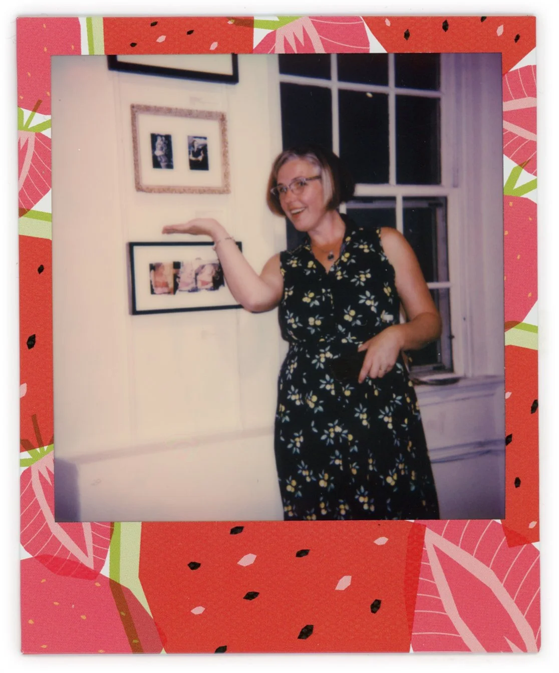 A woman with short hair and glasses, wearing a black sleeveless dress with a yellow floral pattern, standing in front of a wall with framed pictures, smiling and gesturing towards the pictures, with a window behind her.
