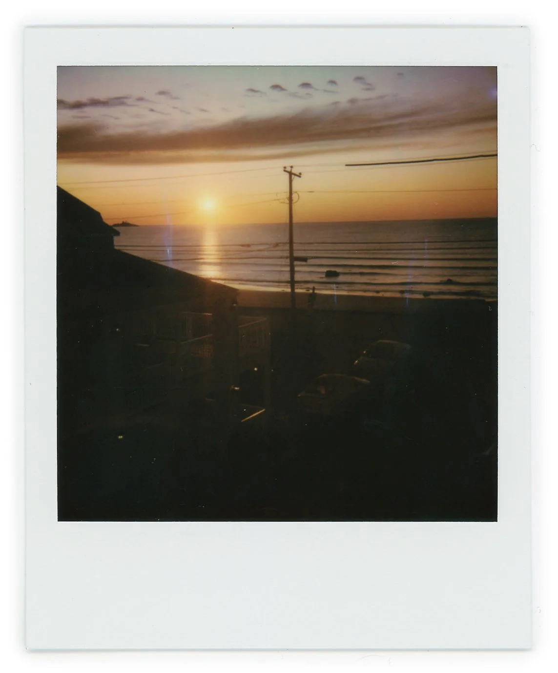 Sunset over the ocean with a silhouetted beachfront, utility poles, and parked cars in the foreground.